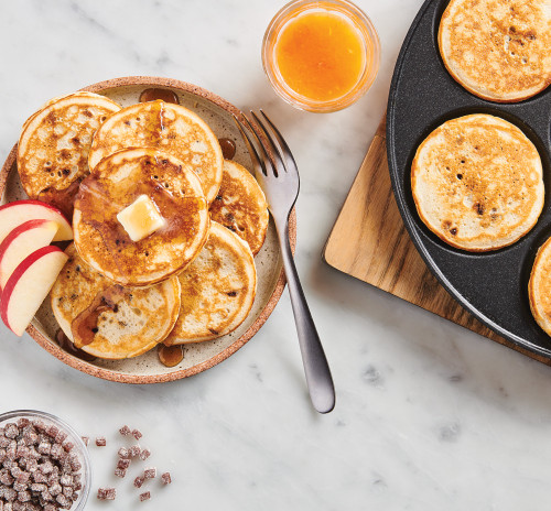 Silver dollar pancakes on a plate with butter and syrup, apple slices, and orange juice in a glass.