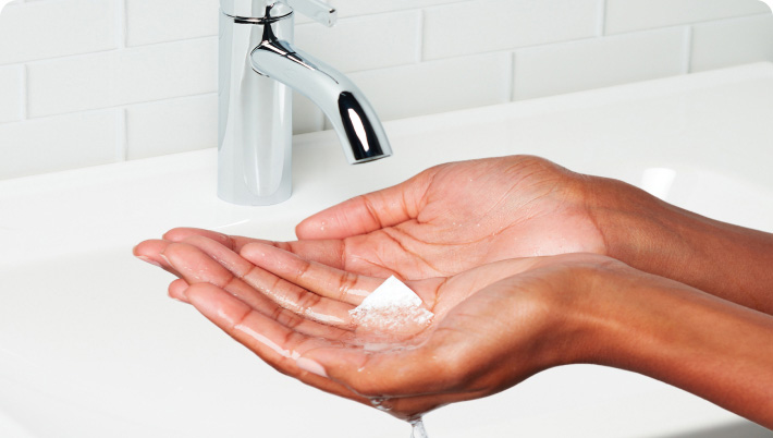 A person washing hands with foamy cleanser under a running faucet in a white tiled bathroom.