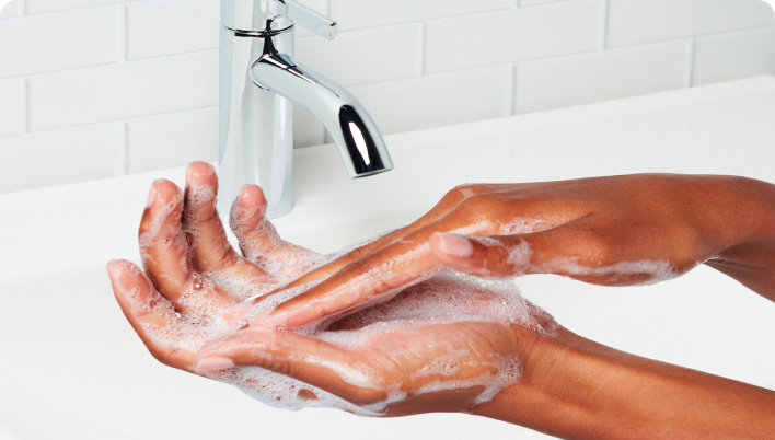 A person rubbing their hands with foamy cleanser under a running faucet in a white tiled bathroom.