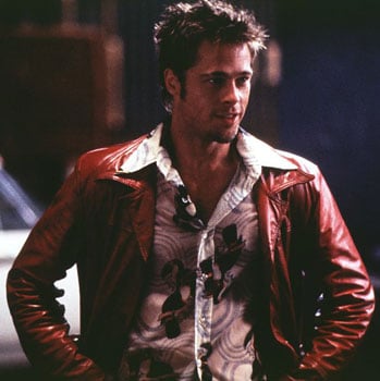 Brad Pitt, wearing a red leather jacket, is standing in front of a car.