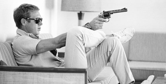 A man sitting on a couch holding a gun, channeling Steve McQueen for style inspiration in khakis.