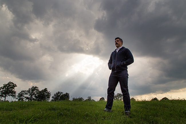 A man in a hoodie standing in a field under a cloudy sky.