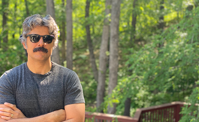 A man with sunglasses standing in a wooded area.