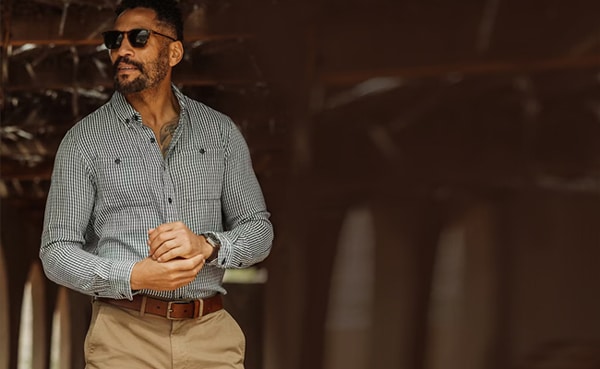 A stylish man in a smart casual checked shirt and sunglasses standing confidently with his sleeves rolled up.