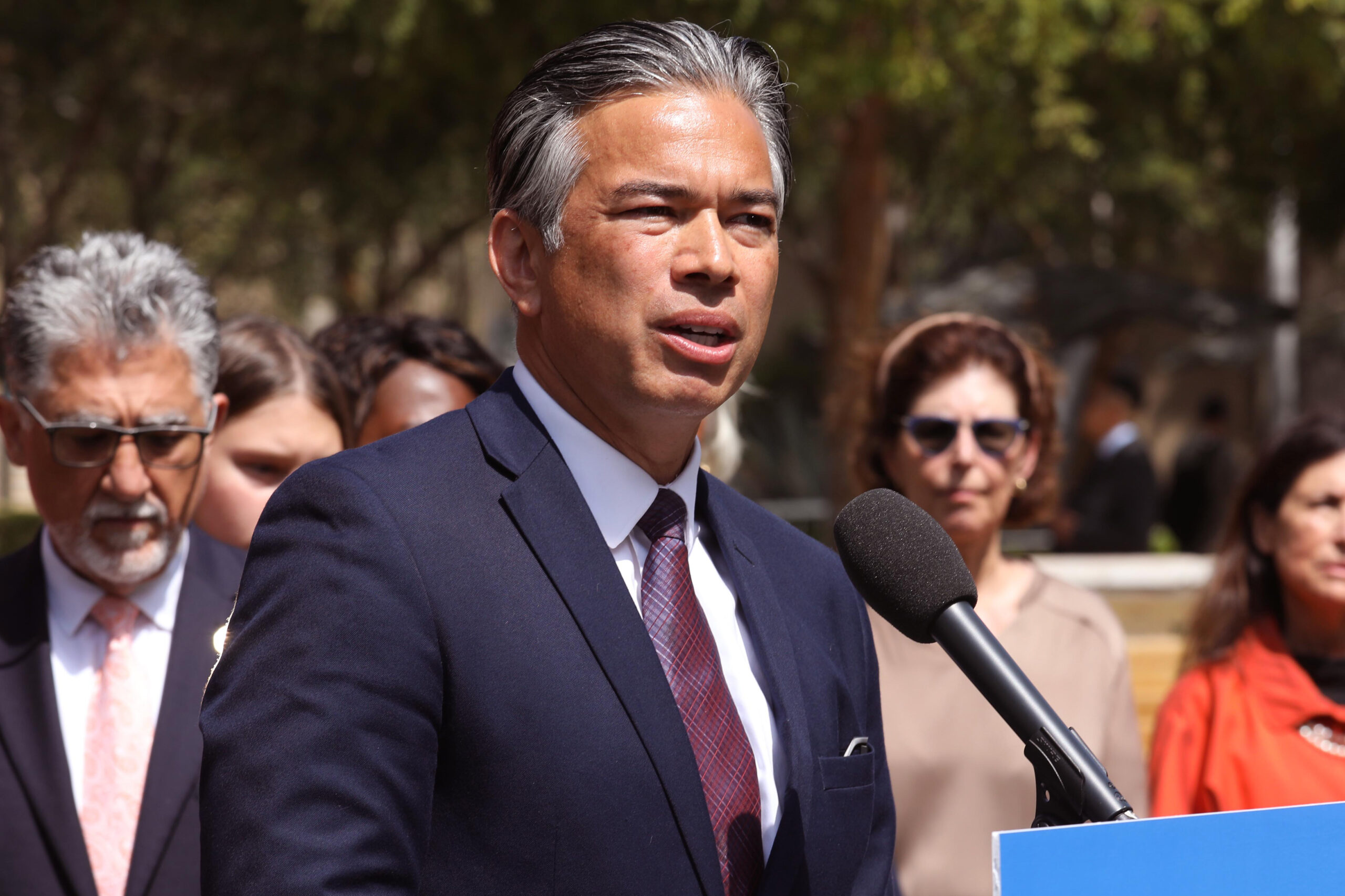 A man in a suit speaks at a podium with a microphone; others listen in the background, outdoors under daylight.