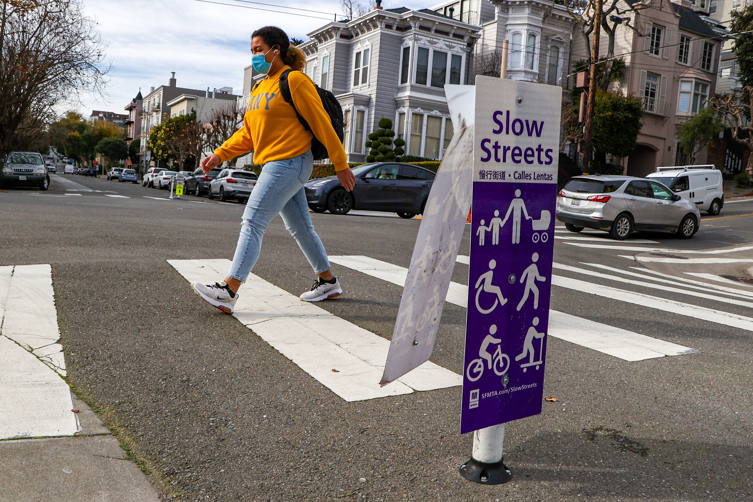 A pedestrian crosses a city street near a "Slow Streets" sign.