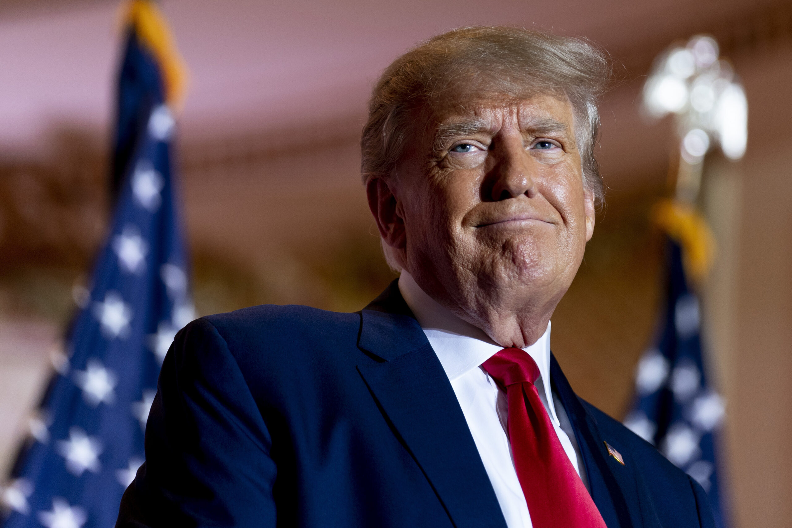 A man with light hair and a suit, featuring a red tie, stands in front of American flags with a composed expression on his face.