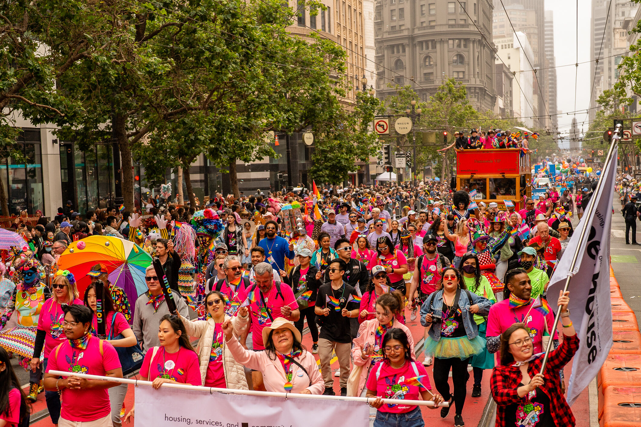 A large, lively crowd participates in a festive parade on a city street. Many wear bright clothing, rainbow accessories, and hold signs and flags, with a double-decker bus in the background.