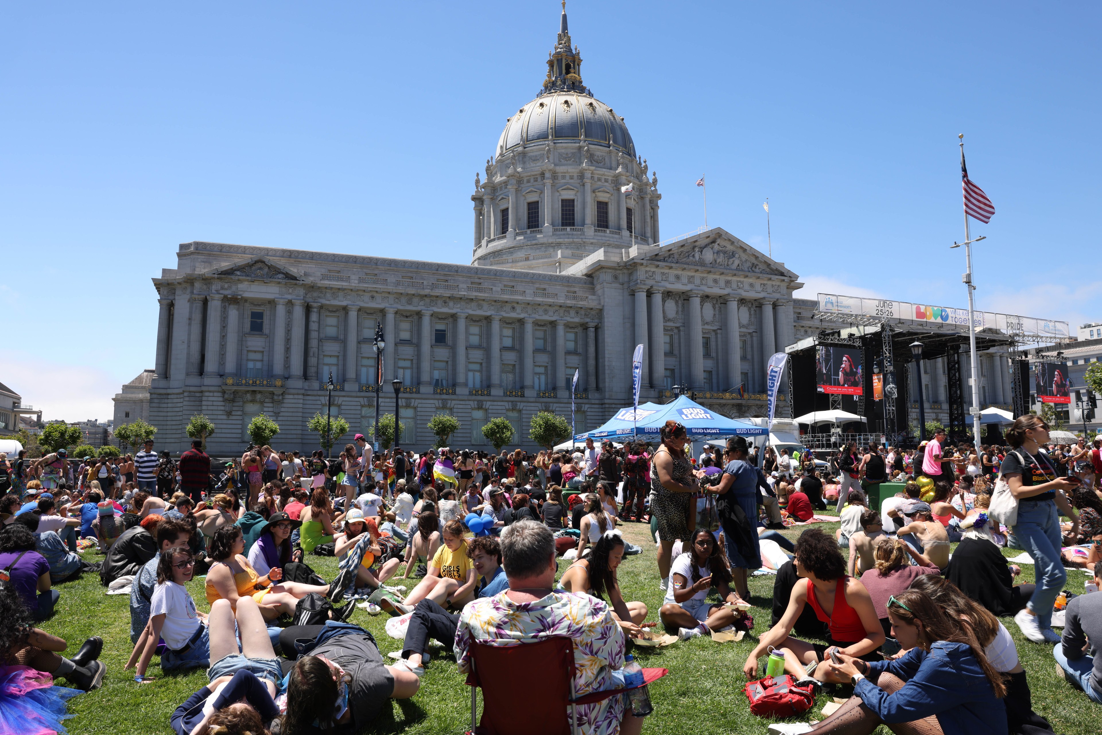 Hundreds of people are gathered on a lawn in front of a large domed building, with a stage and screens visible in the background, during a sunny outdoor event.