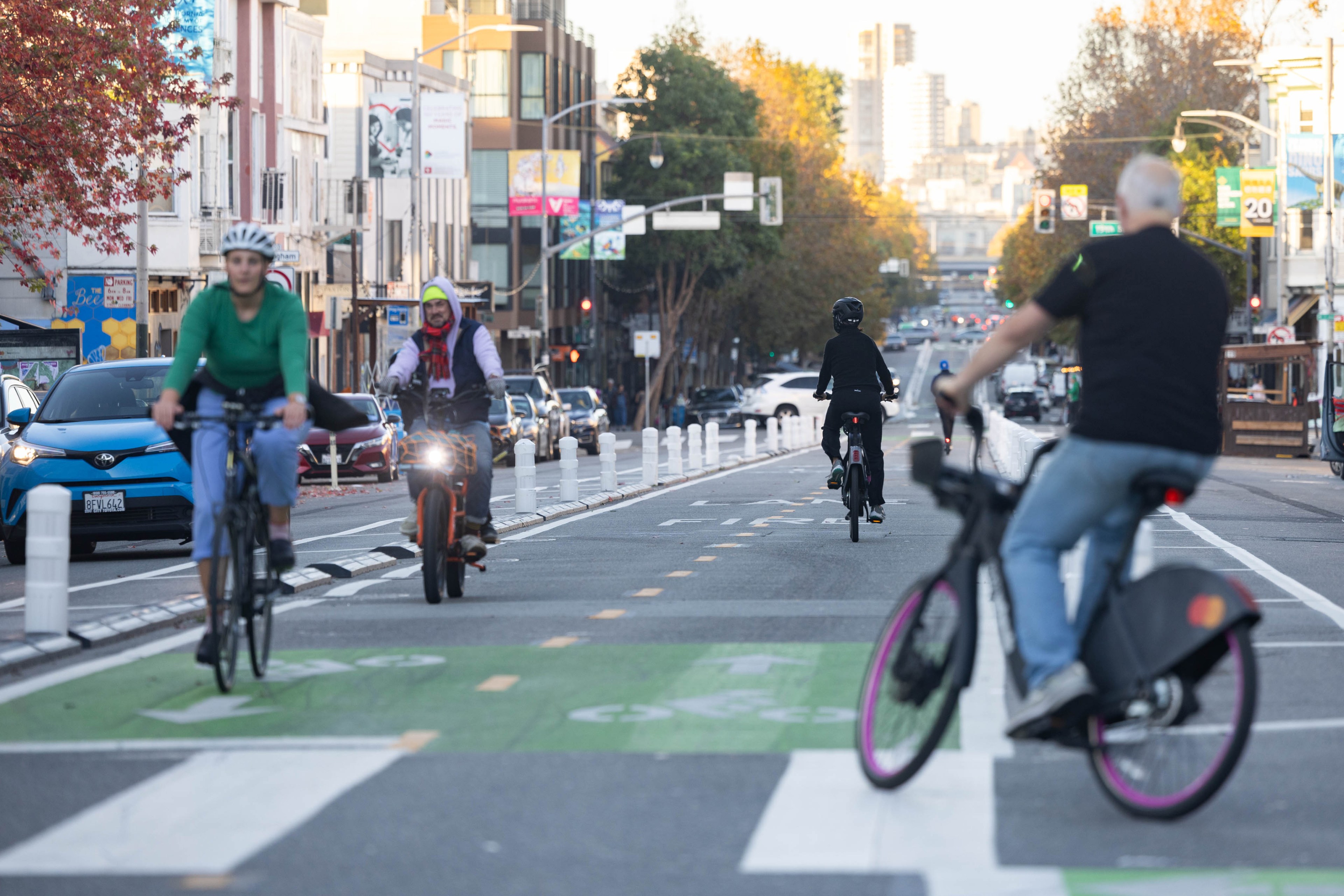 A long view down a street shows cyclists and a scooter rider in a green painted bike lane with a crosswalk in the foreground.