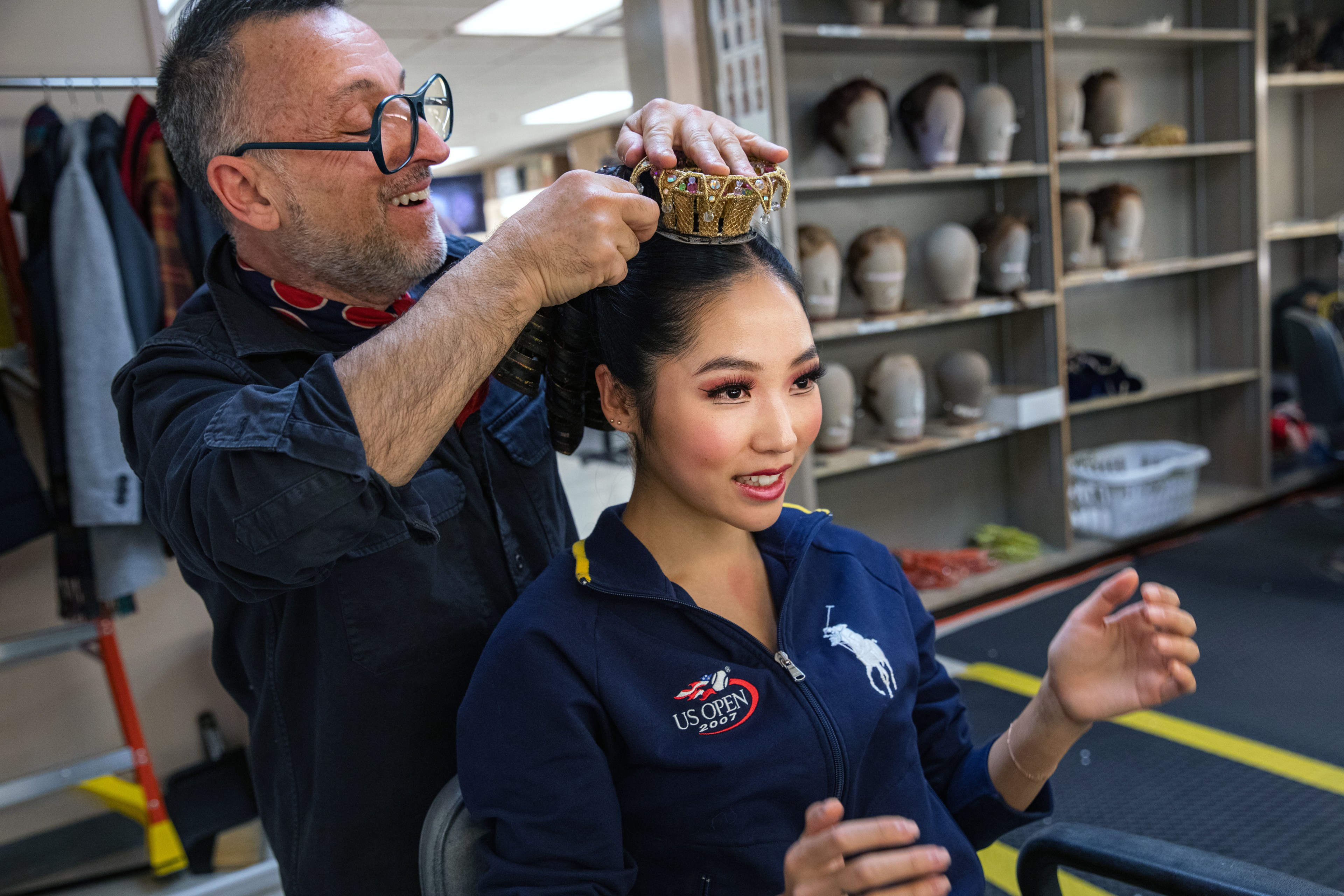 A woman has a small crown put on her head while backstage with mannequin heads in the background.