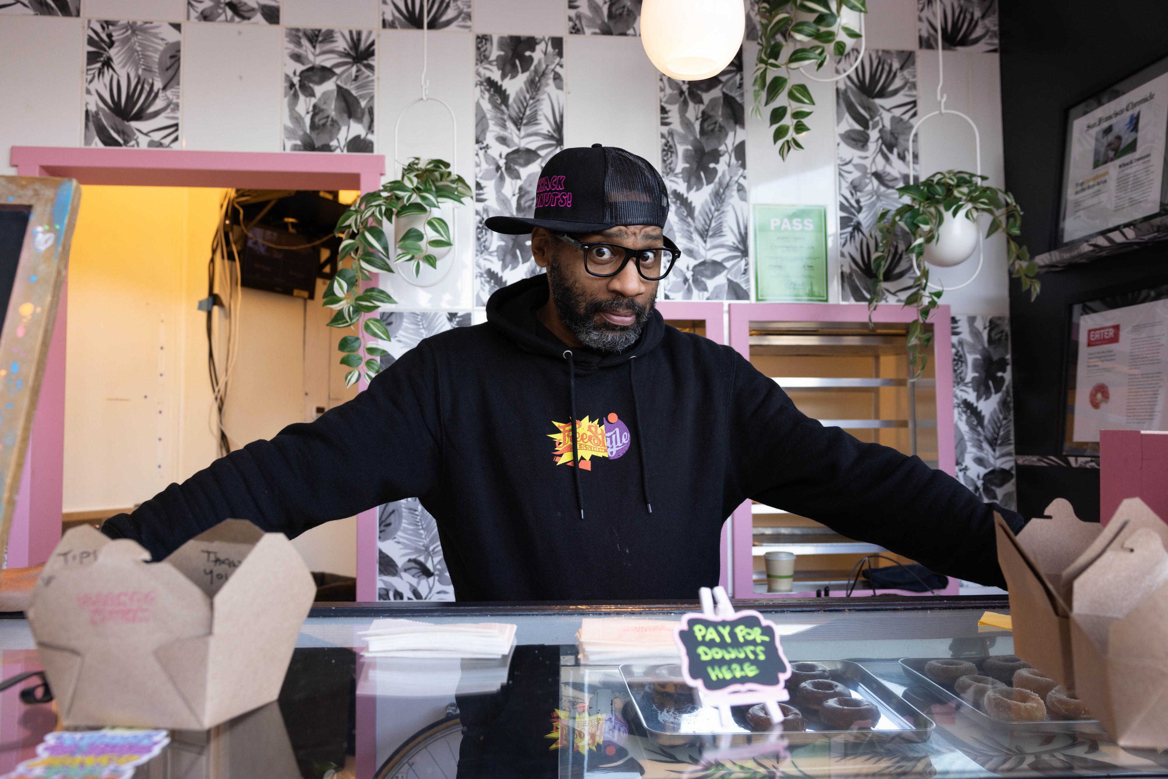 A man, Vandor Hill, owner of Whack Donuts, poses for a portrait while standing behind a counter filled with donuts.