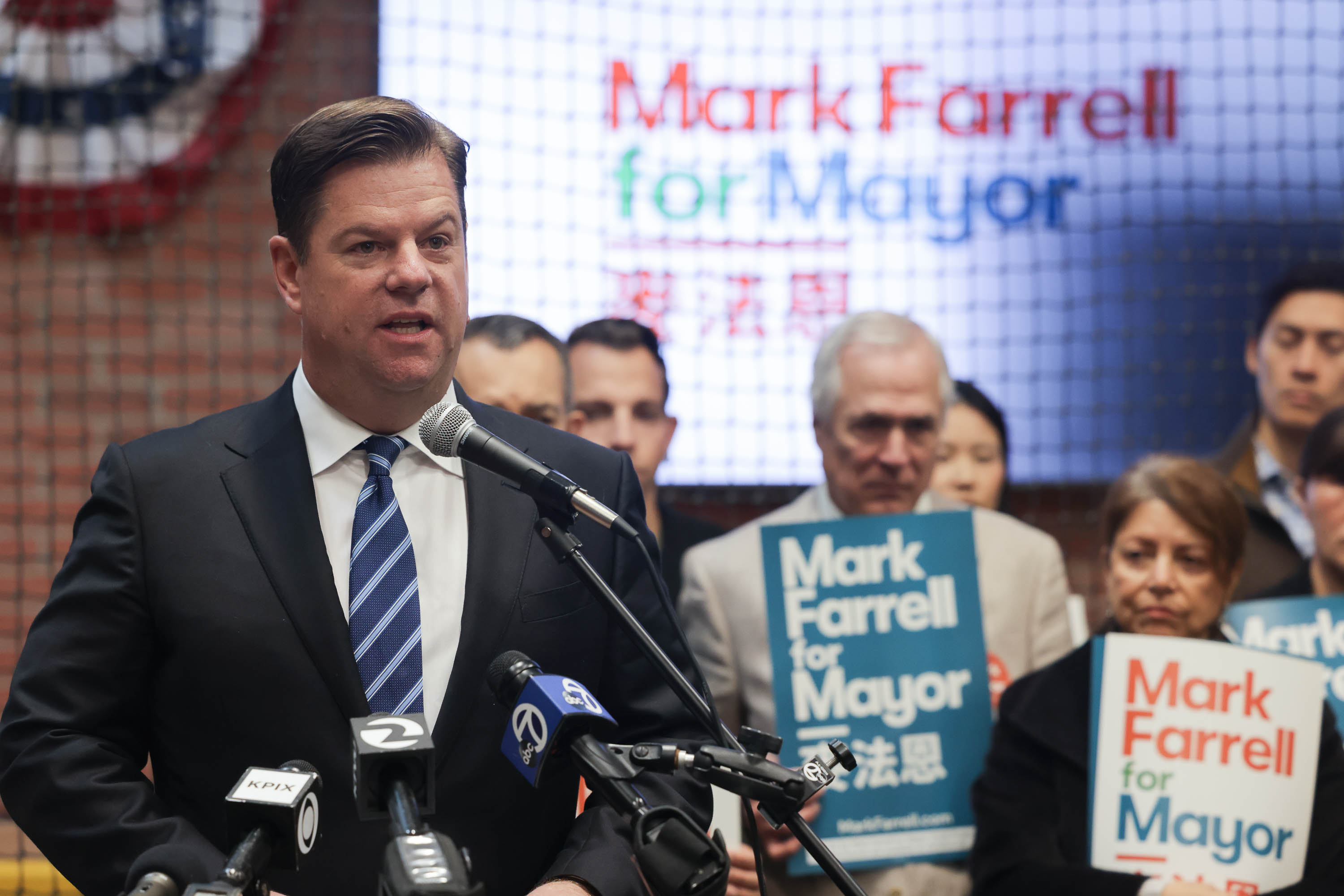 A man at a podium with a &quot;Mark Farrell for Mayor&quot; sign and supporters behind him.