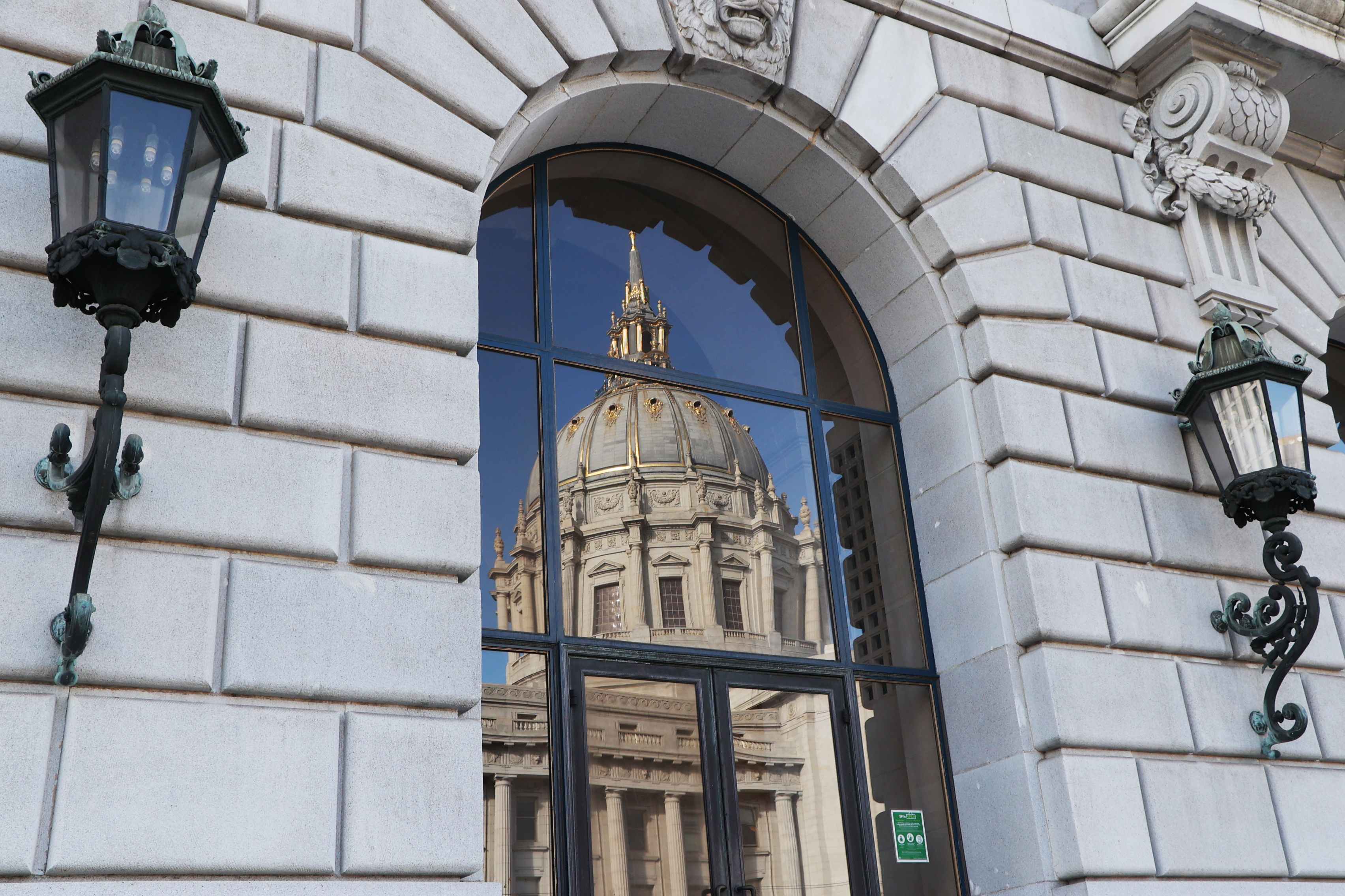 The image shows a domed building reflected in a large arched window with ornate, black street lamps on either side, set against a gray stone facade.