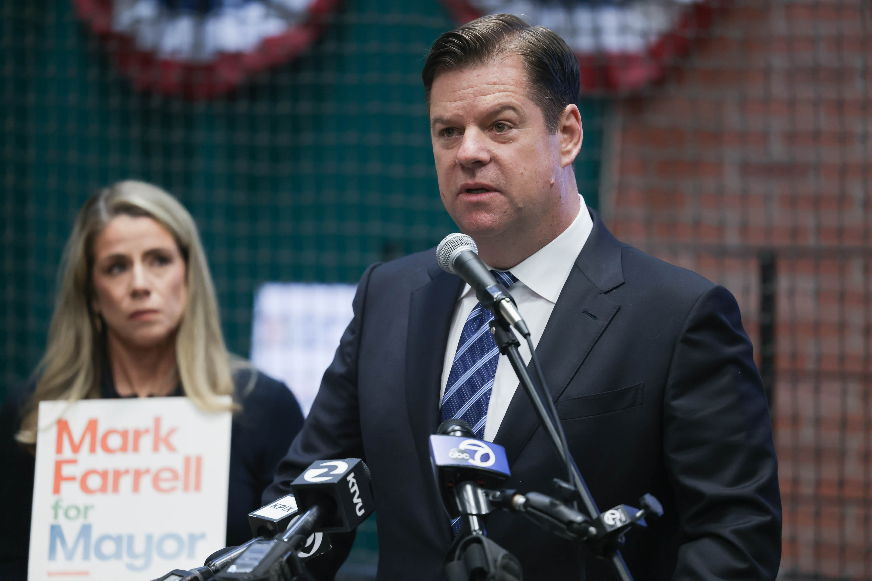 A man speaks into microphones at a podium with a campaign sign, while a focused woman listens behind him.