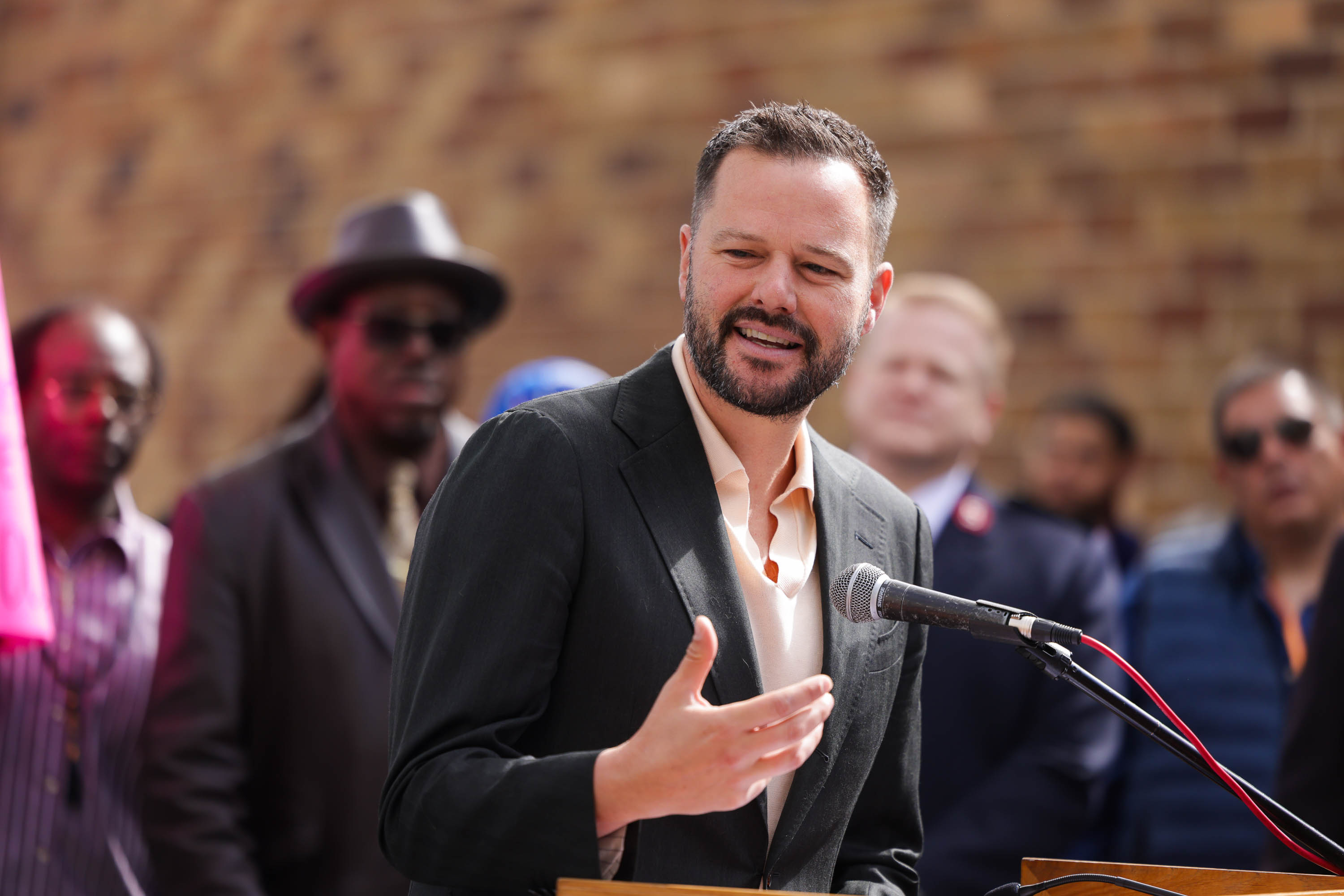 Matt Haney in a suit is speaking at a podium with a microphone, gesturing with his hand. Several people are standing behind him against a blurred brick background.