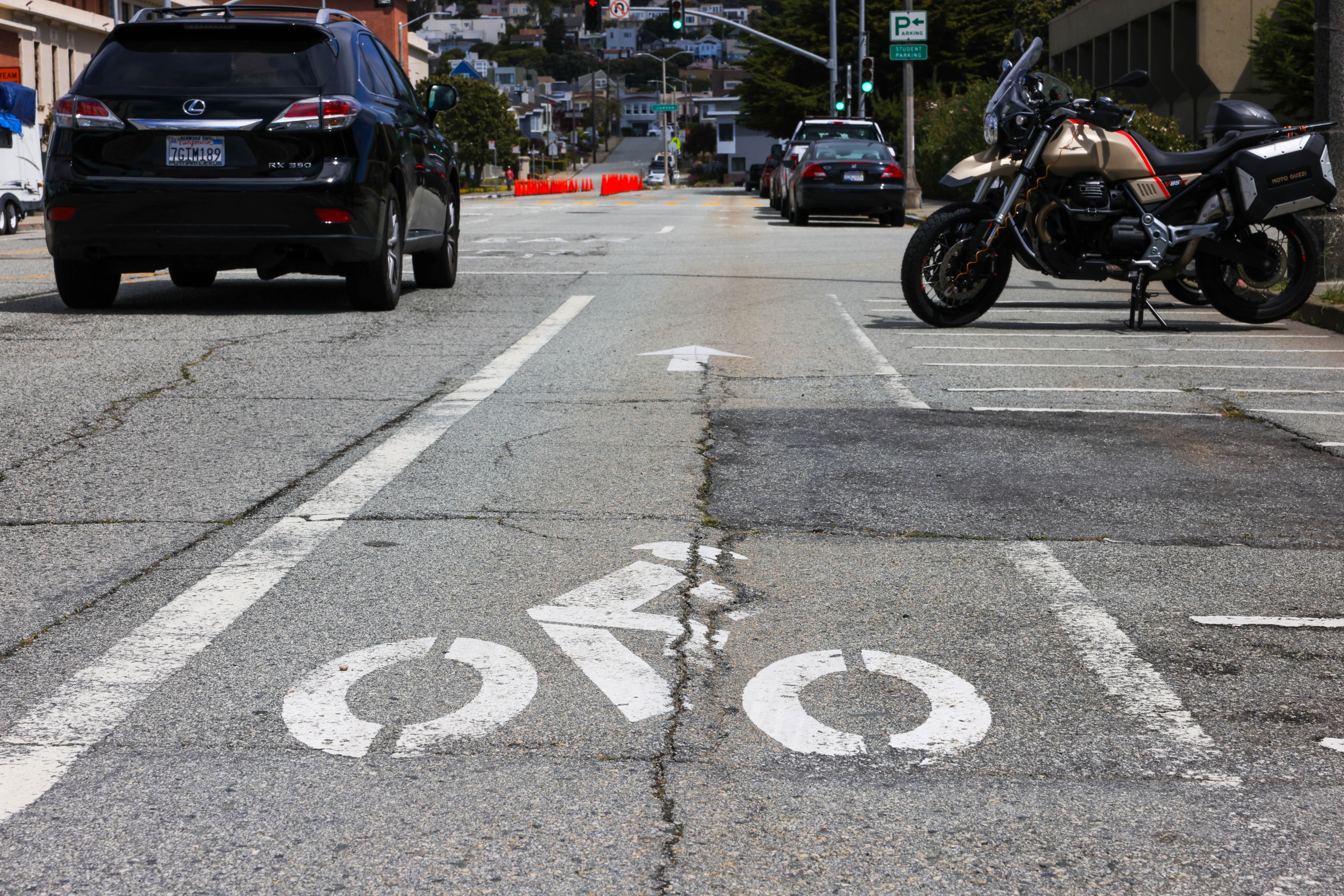 A city street with a bike lane symbol, cars on the road, and a parked motorcycle to the right.