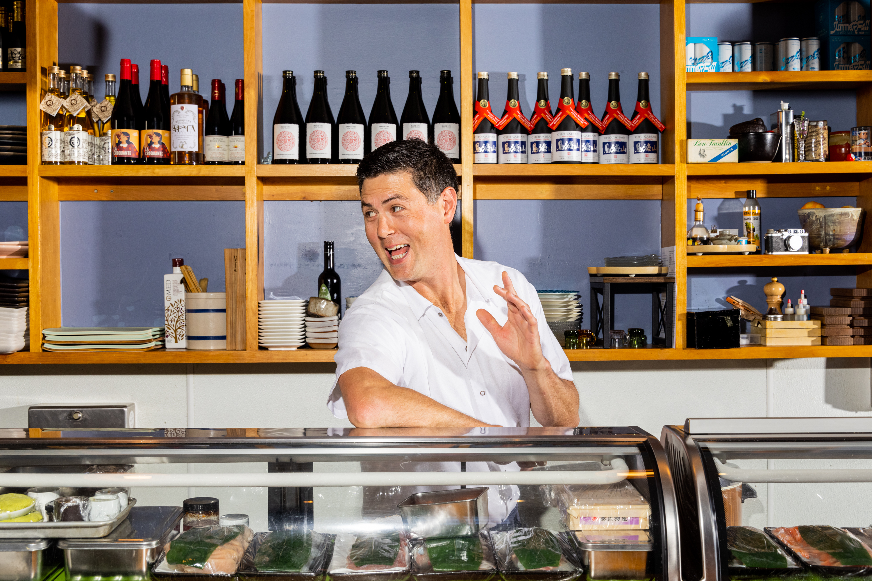 A man in a white shirt stands behind a counter in front of shelves stocked with bottles and dishes, smiling and gesturing with his right hand raised.