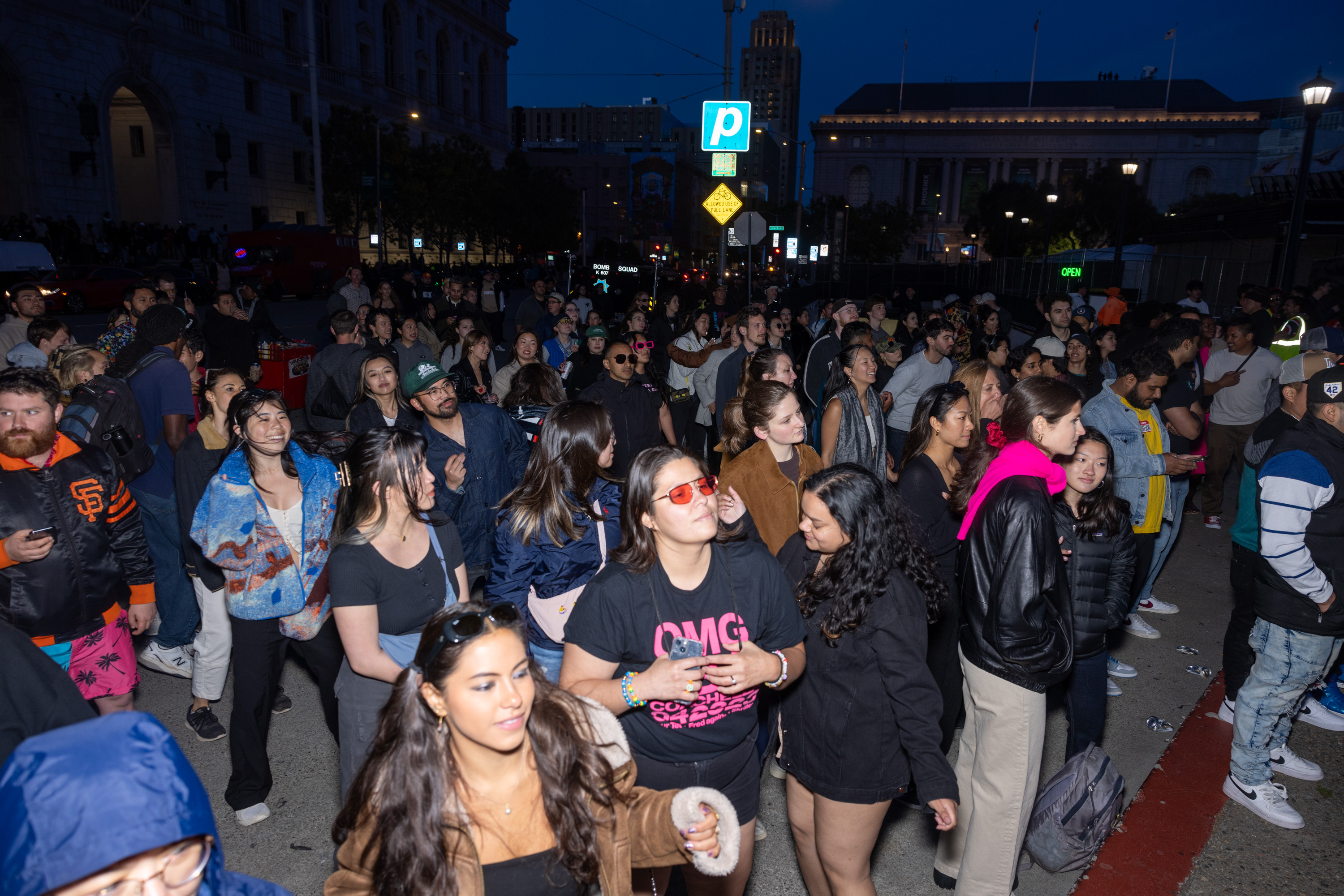 A large, diverse group of people is gathered outdoors at night. Most are standing and appear to be enjoying an event, with some smiling and chatting. Buildings and street signs are visible in the background.