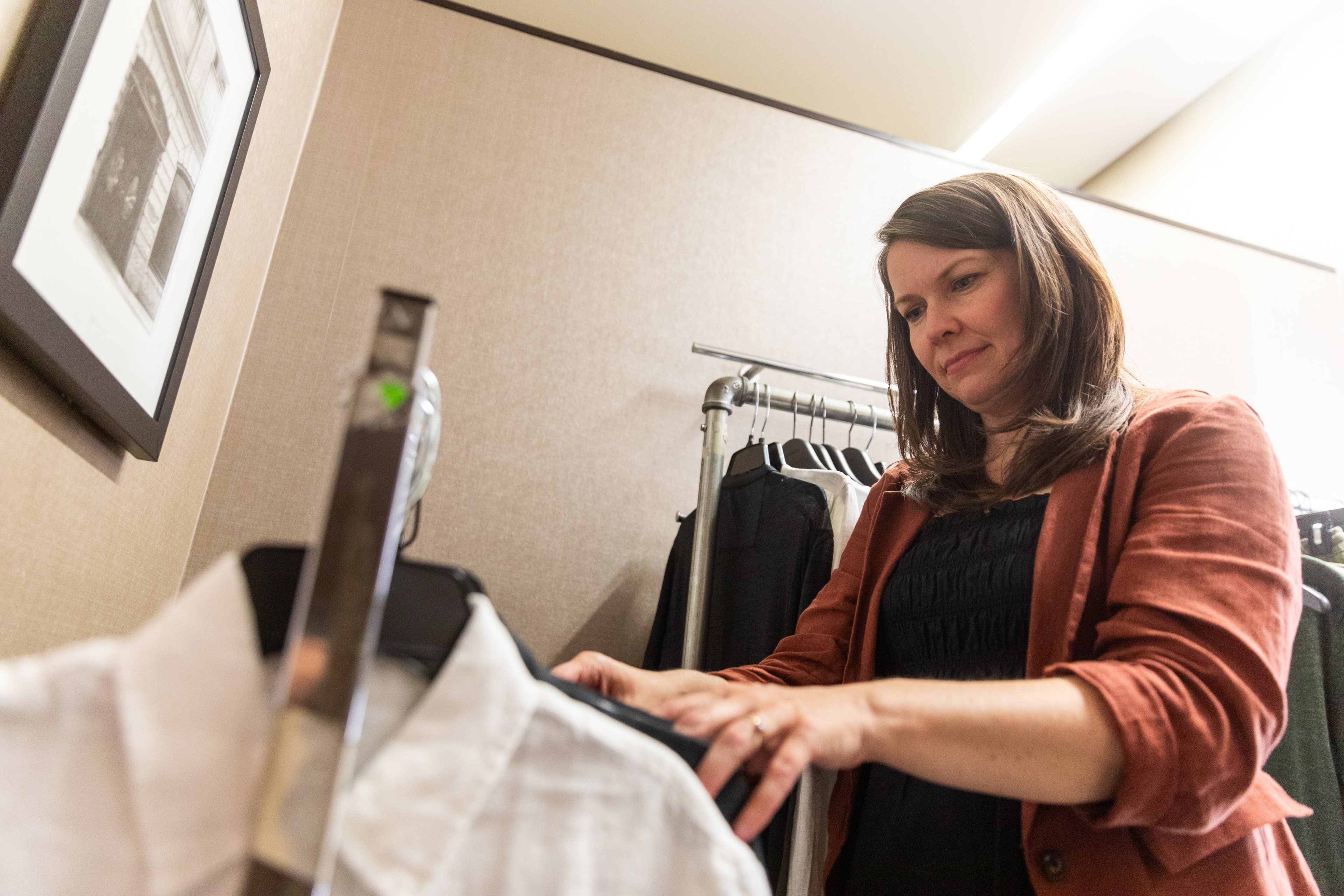 A woman in a rust-colored blazer browses clothing on a rack in a well-lit room with beige walls and framed artwork.