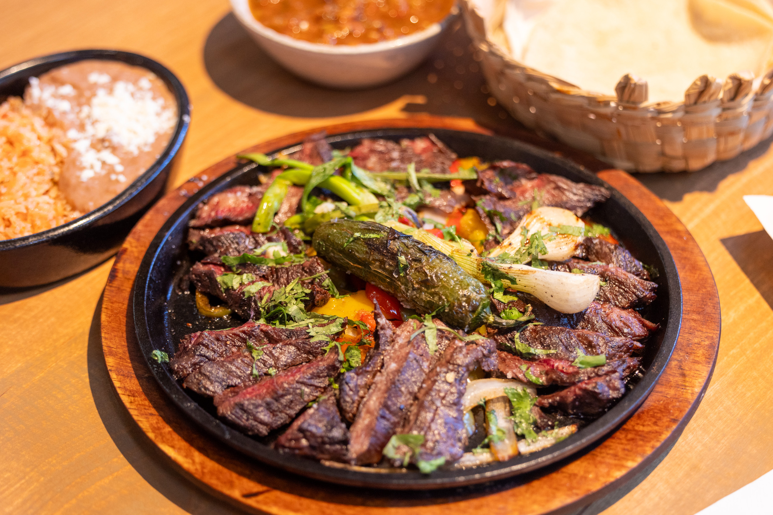 A cast iron skillet with steak, onions, and peppers sits on a wooden table. In the background: a plate of rice and beans, salsa, and toritllas.