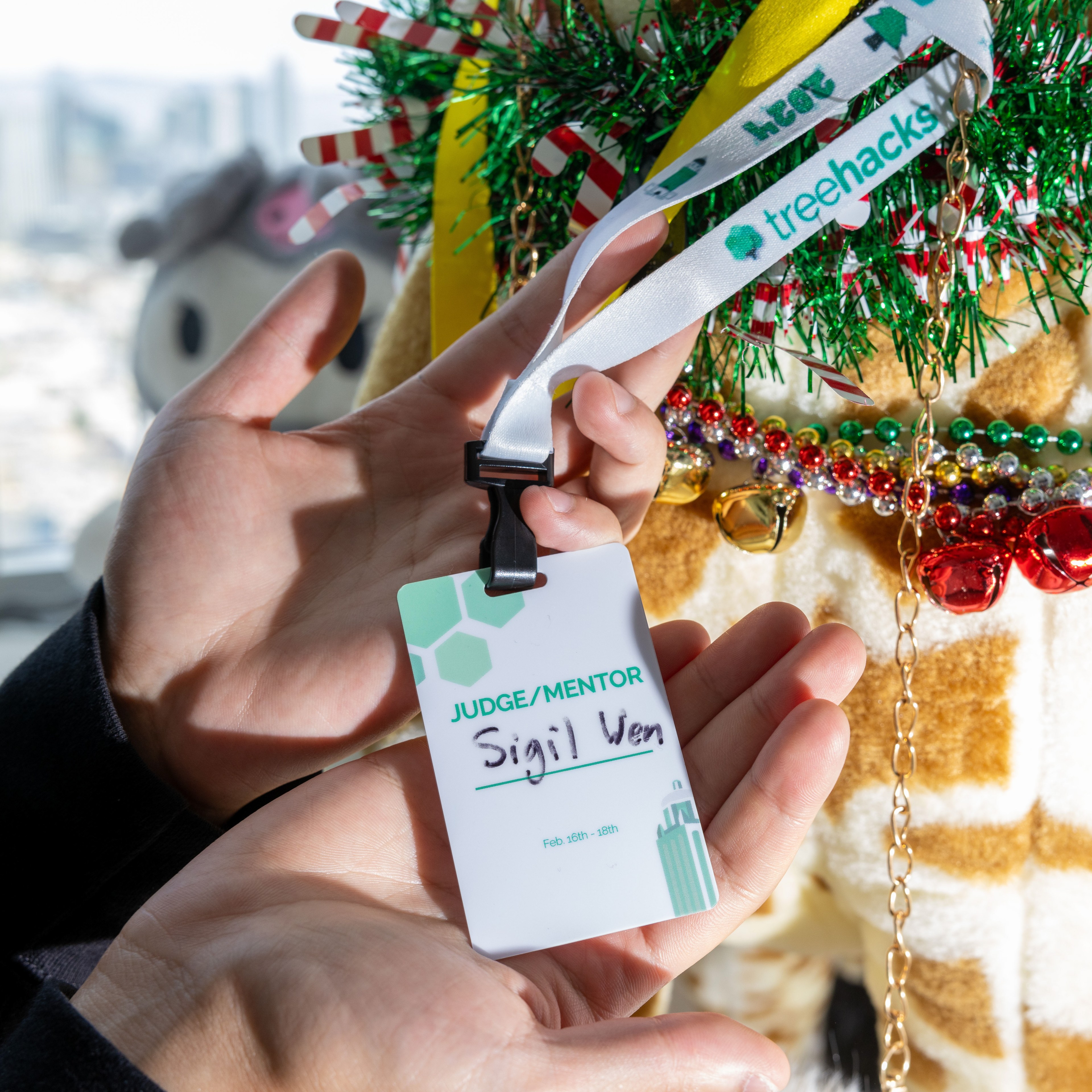 The image shows a person holding a name badge that reads &quot;Judge/Mentor&quot; and &quot;Sigil Ven,&quot; with a lanyard labeled &quot;treehacks.&quot; It appears festive with decorations like beads, candy canes, and garlands in the background.