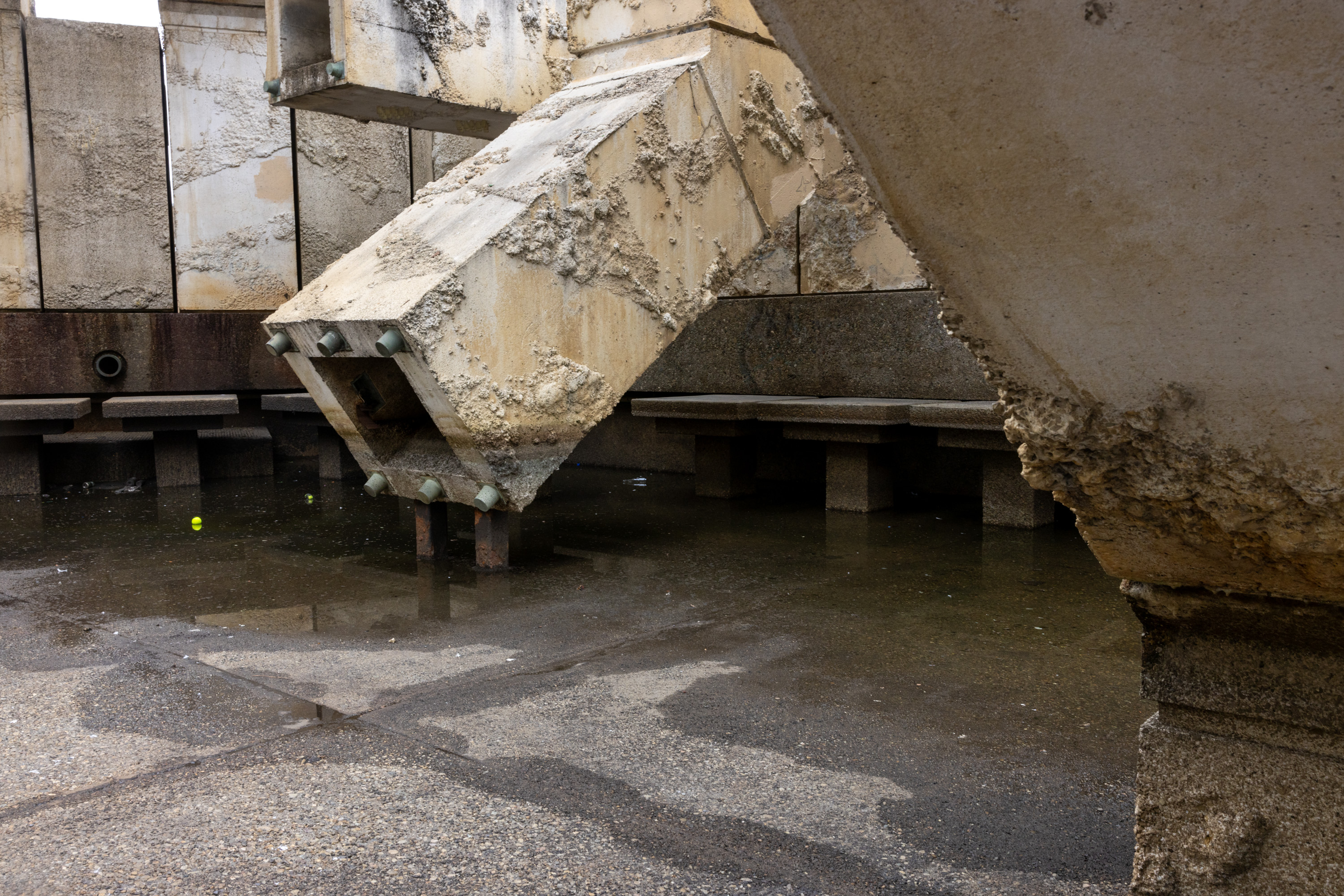 A rugged, weathered concrete structure over a reflective, wet ground with benches in the background. A small yellow ball is visible on the ground.