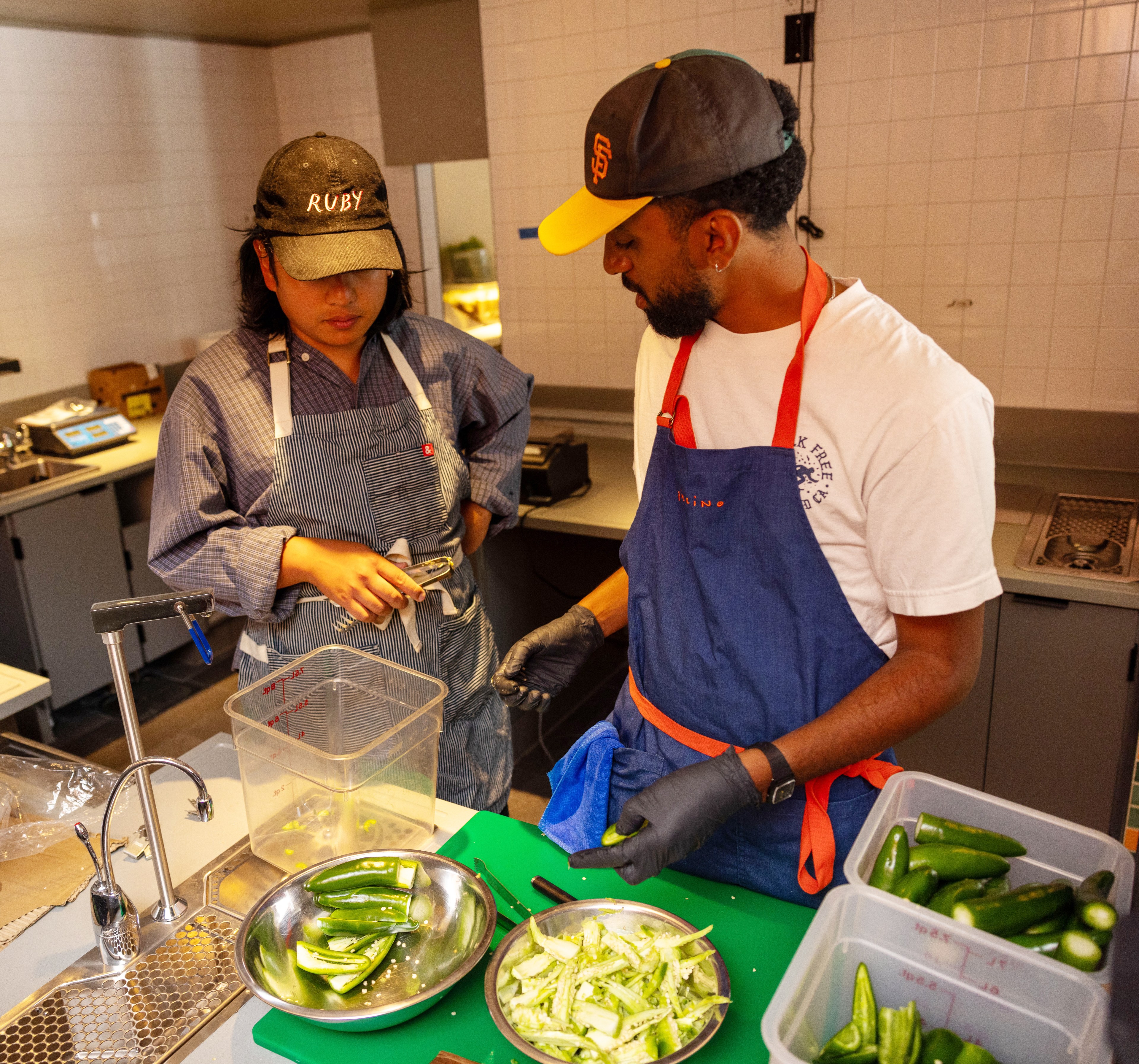 Two individuals are in a kitchen, both wearing aprons and caps. One is holding tongs, while the other is slicing green peppers on a cutting board. Bowls of sliced peppers are on the counter.