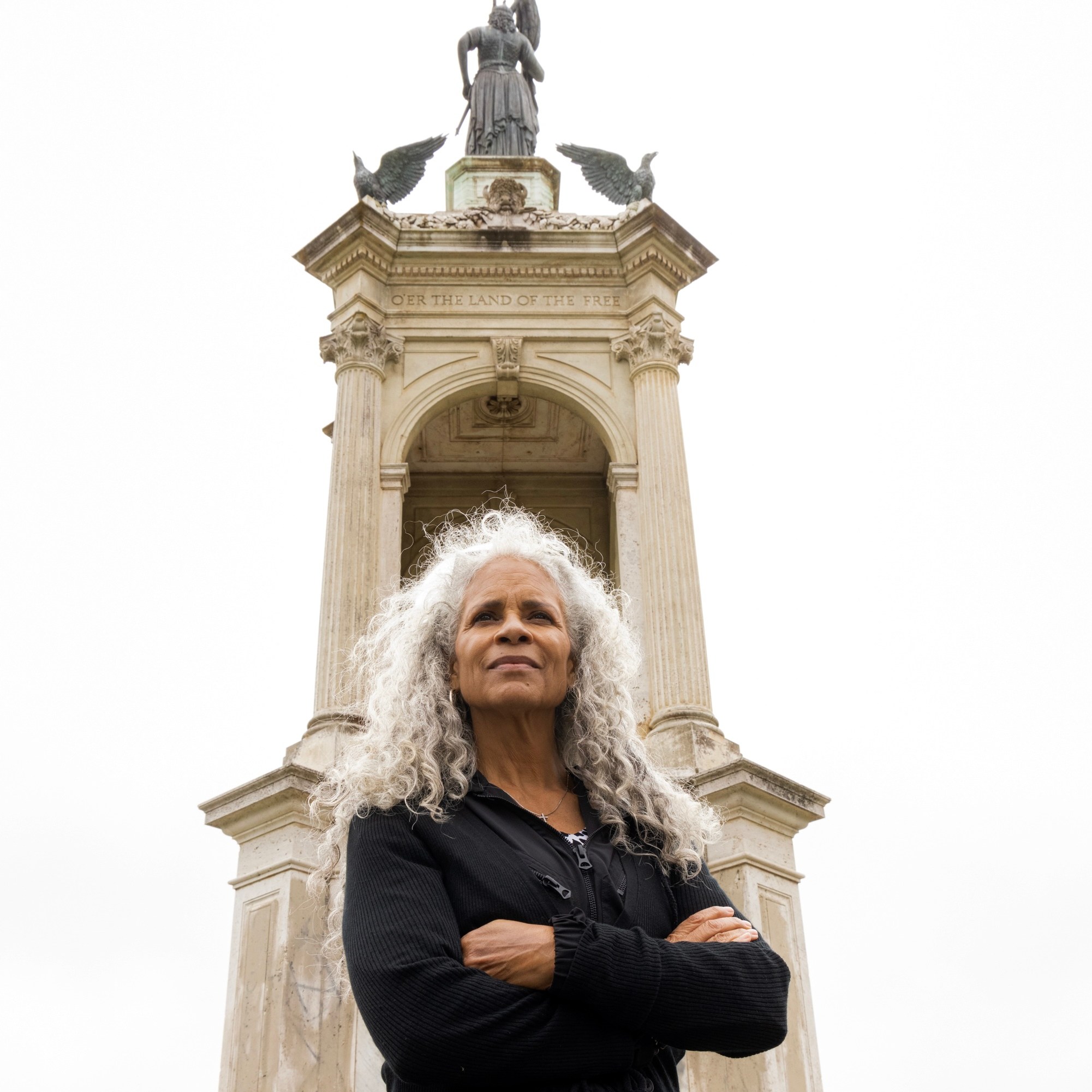 A woman with curly white hair stands confidently with folded arms in front of a tall monument featuring statues and inscriptions against a cloudy sky.