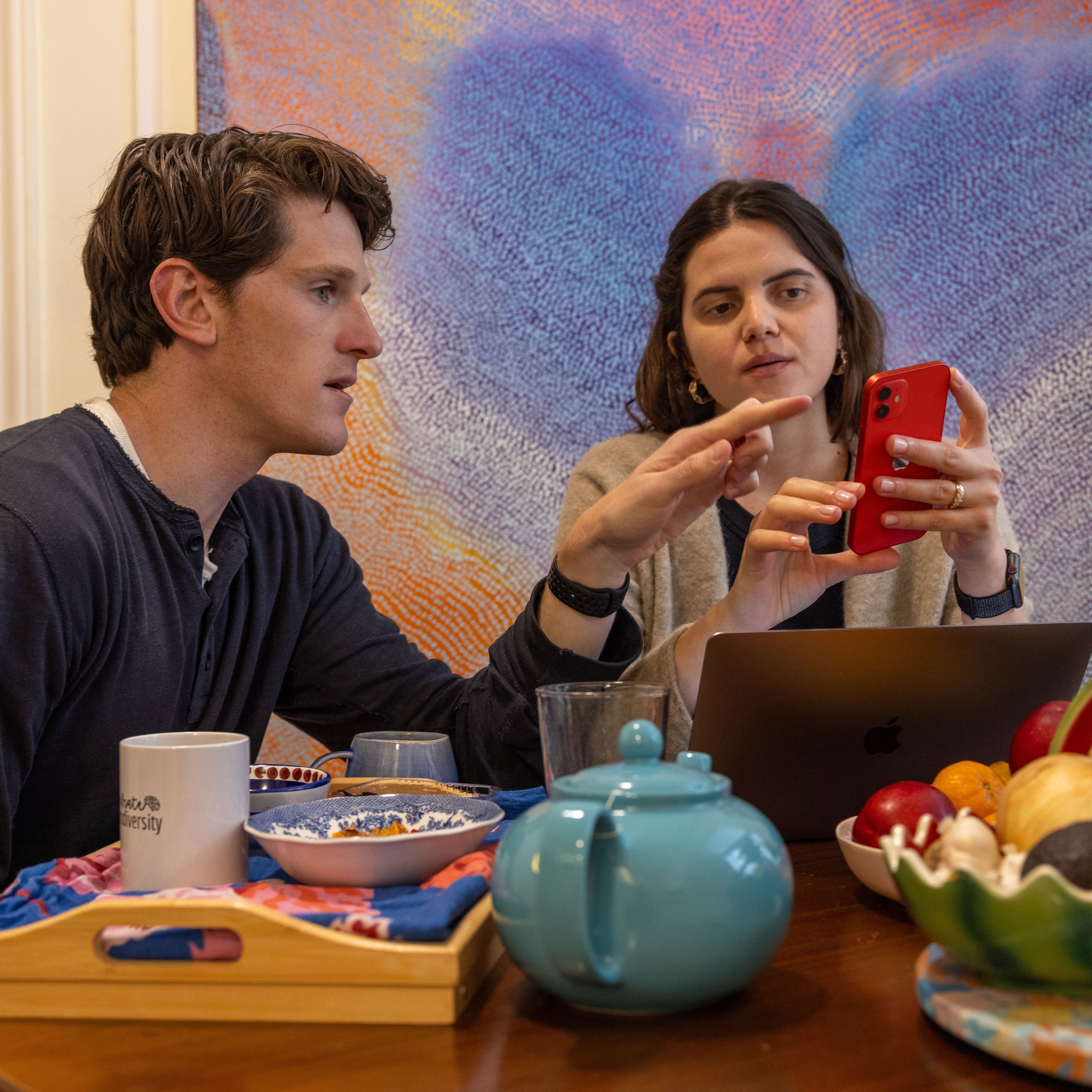 Two people are seated at a table with a colorful background, looking at a red phone. The table has a blue teapot, a tray with food, a mug, a laptop, and fruit.