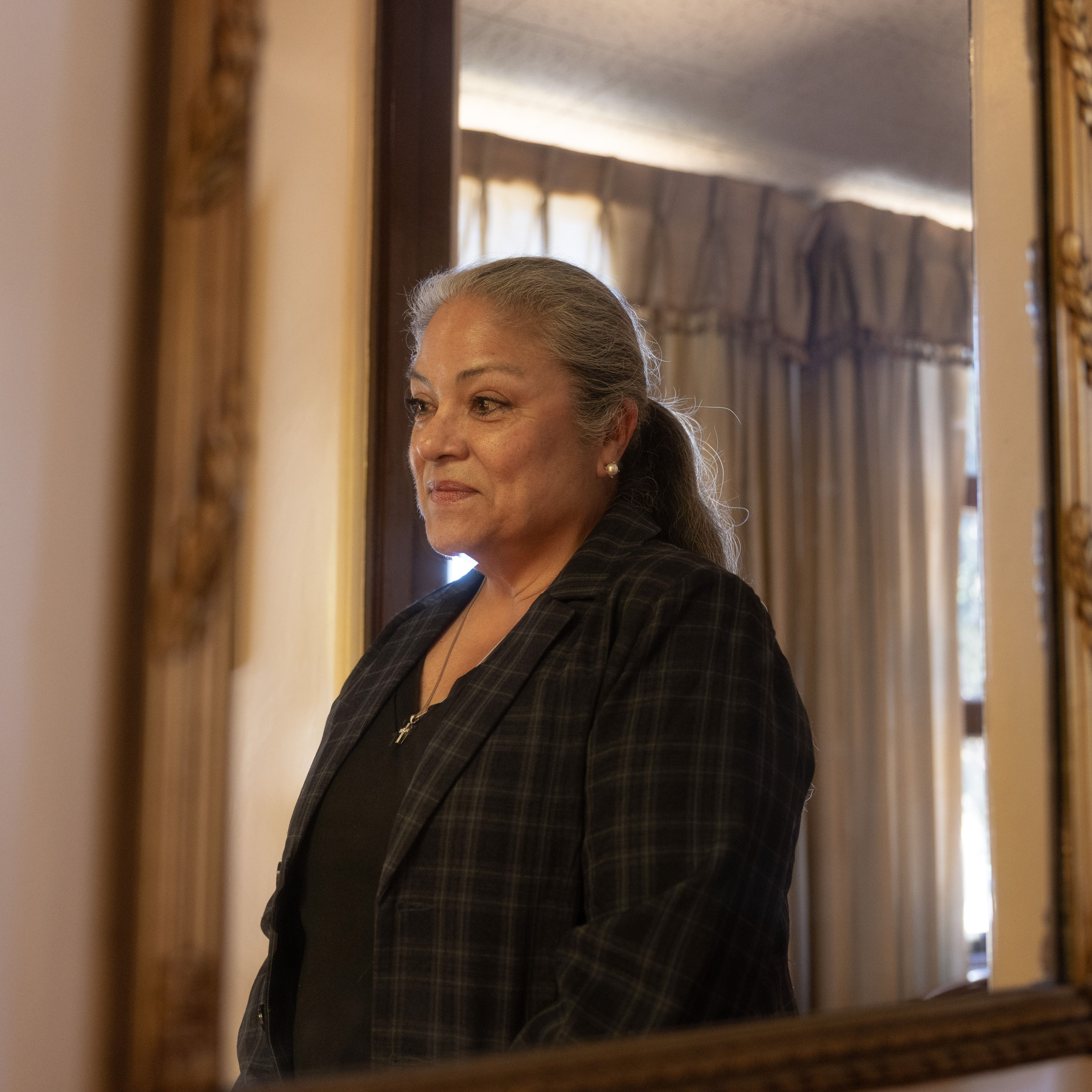 A woman with gray hair in a ponytail, wearing a dark plaid blazer and a necklace, is standing indoors by a mirror with her reflection visible, surrounded by beige curtains.