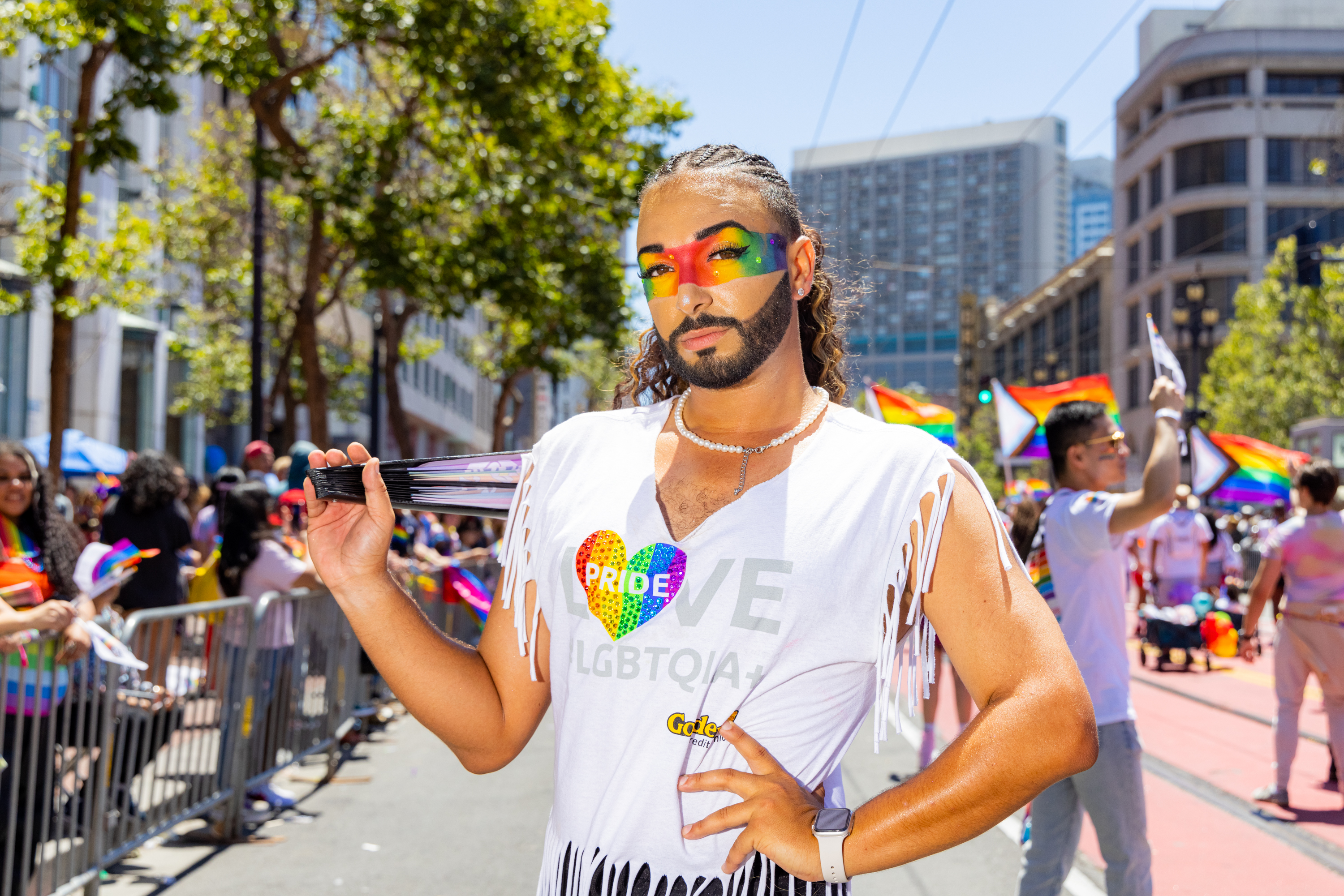 A person with rainbow face paint and a fringed white shirt that says &quot;PRIDE&quot; stands outdoors, holding a fan, among a crowd with rainbow flags and trees in the background.