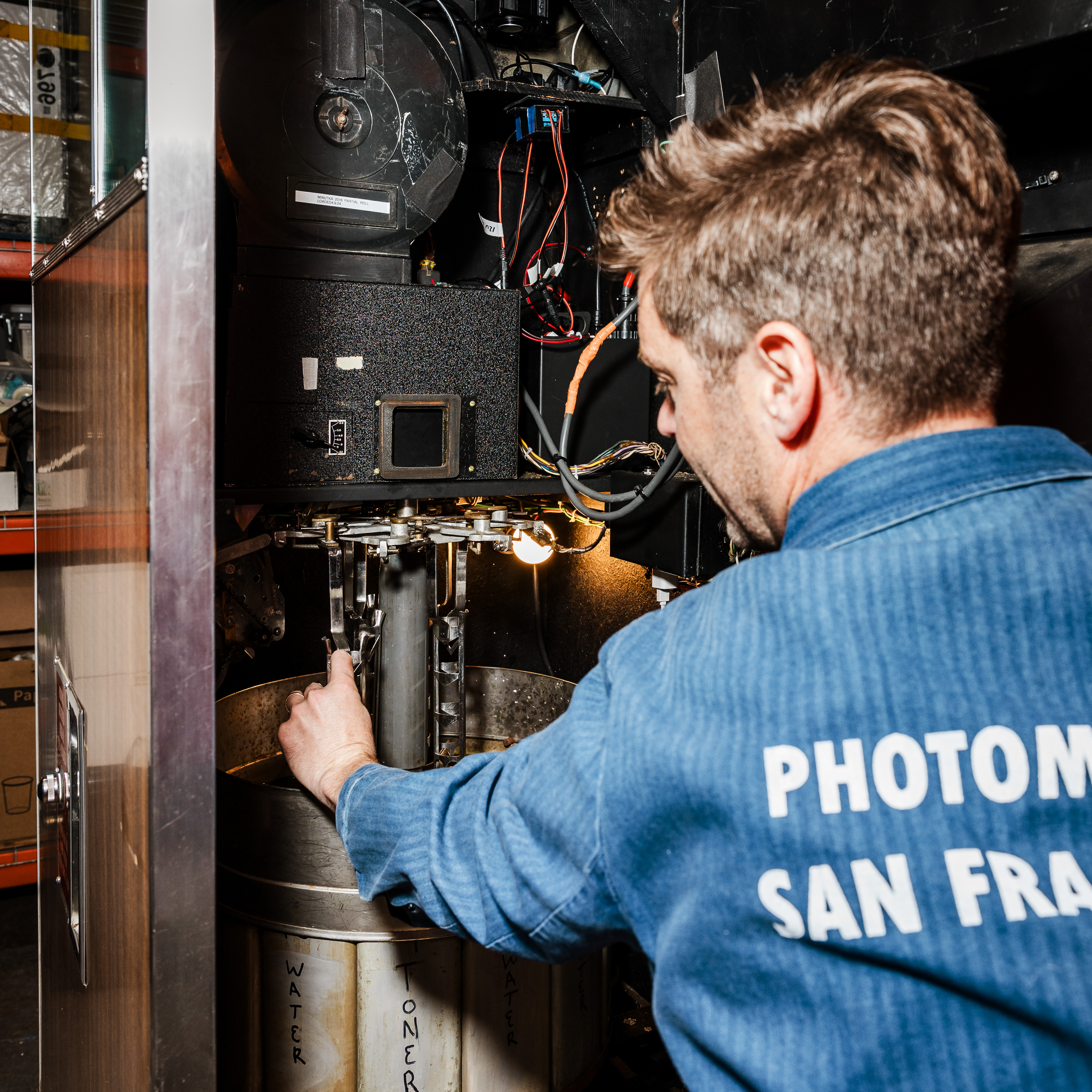 A man in a blue shirt, with &quot;PHOTOM SAN FR&quot; on the back, operates complex machinery, adjusting a component inside while various wires and parts are visible.