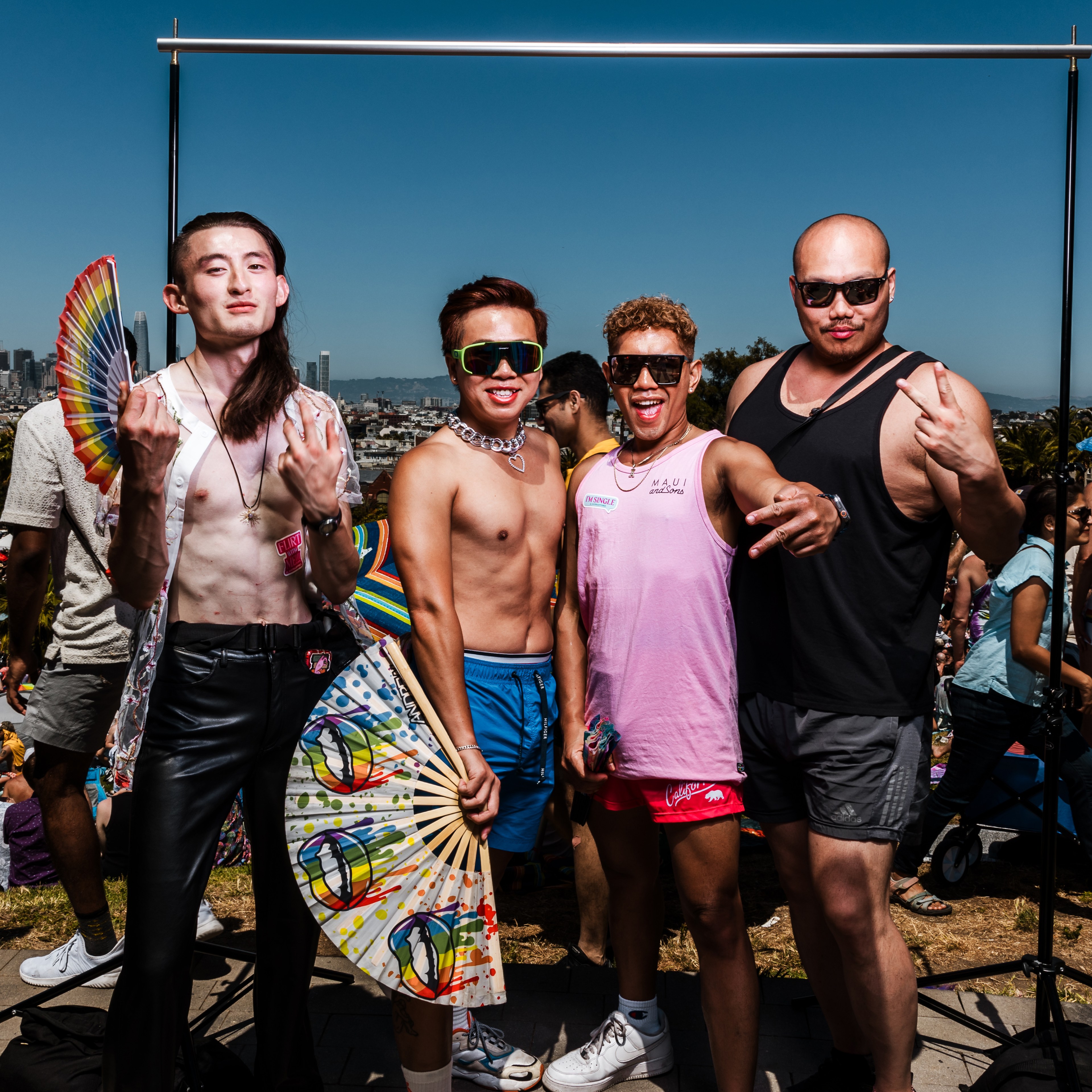 Four people, vibrantly dressed and smiling, pose for a photo outdoors. They hold colorful fans and make peace signs, with a clear blue sky and cityscape in the background.