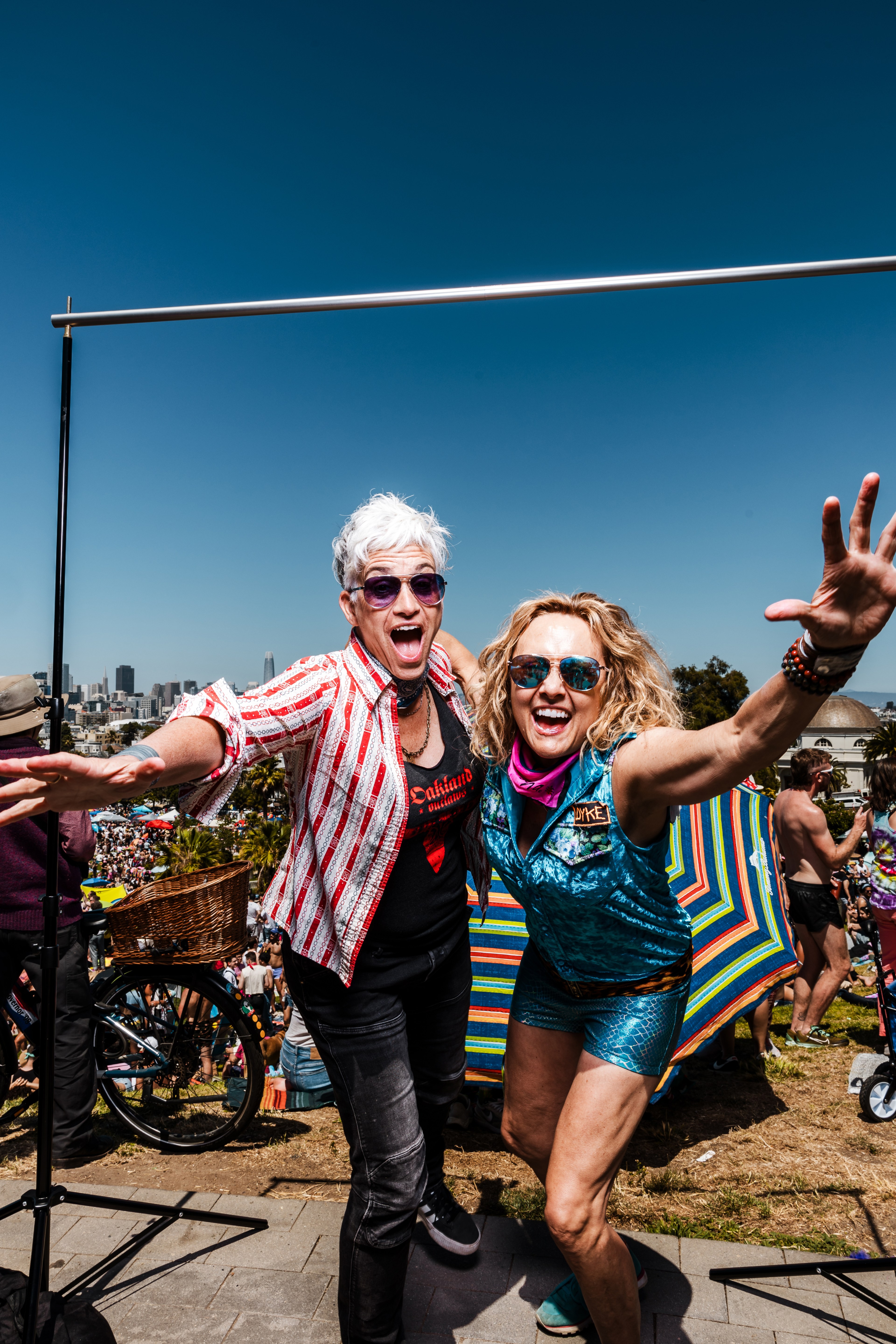 Two people with outstretched arms in colorful outfits and sunglasses are excitedly posing at an outdoor event, with a crowd, bicycles, and umbrellas in the background.