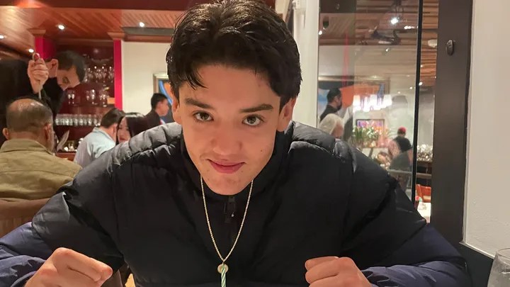 A young person with dark hair is sitting at a table in a restaurant, smiling slightly, and leaning forward with fists on the table. People and soft lighting are in the background.