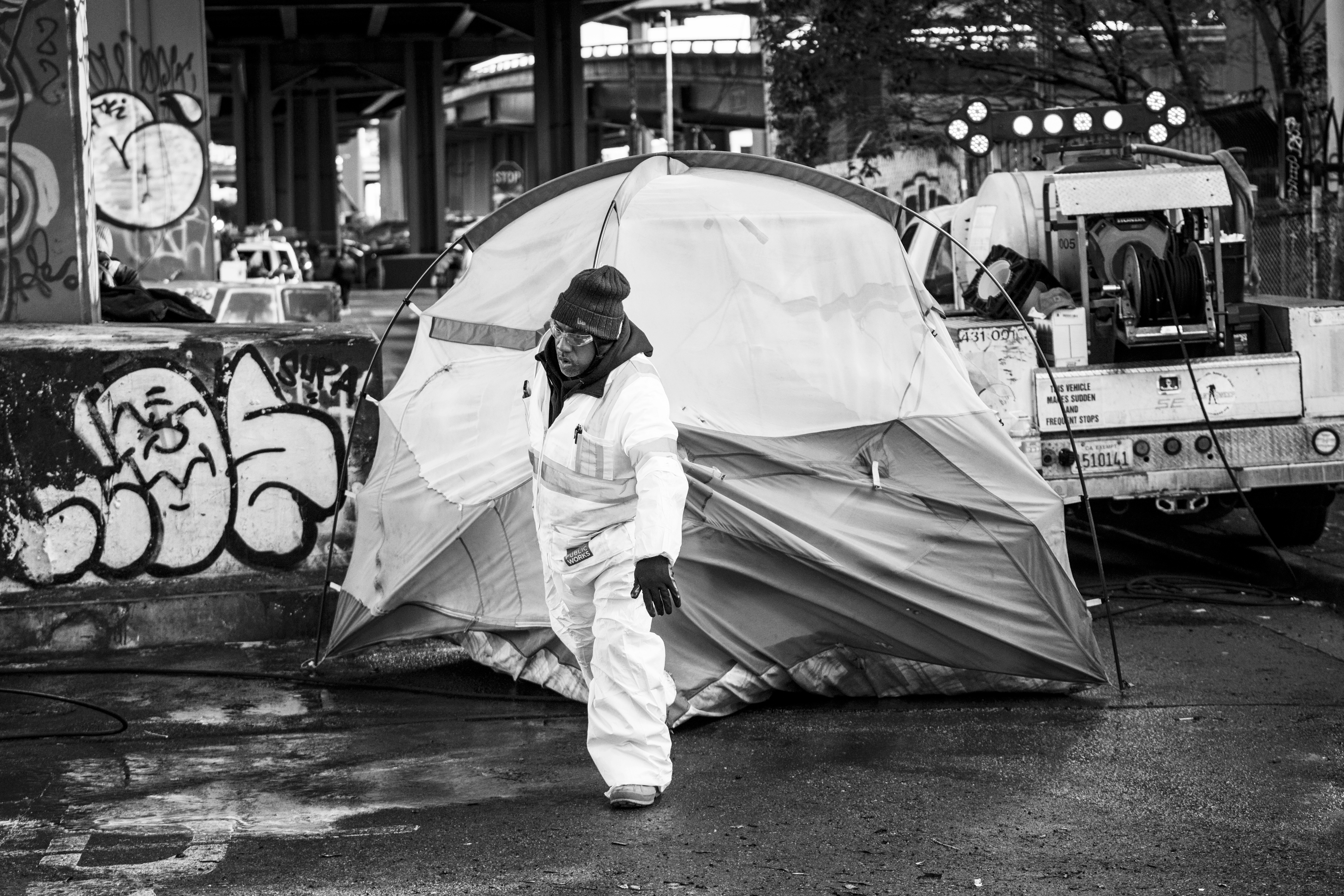 A person in a protective suit walks past a partially collapsed tent under a highway overpass, with graffiti on a concrete barrier and industrial equipment nearby.