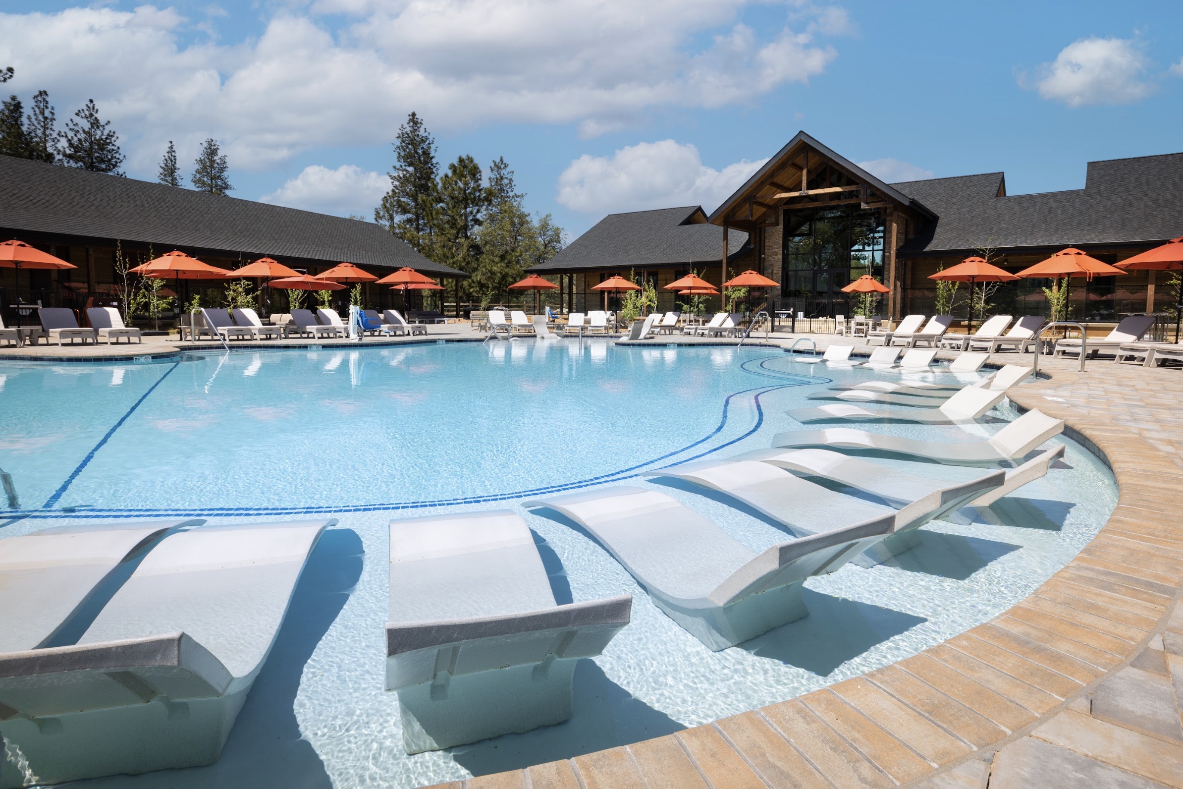 A clear, inviting pool surrounded by lounge chairs and orange umbrellas, set against a backdrop of buildings and trees under a sunny sky.