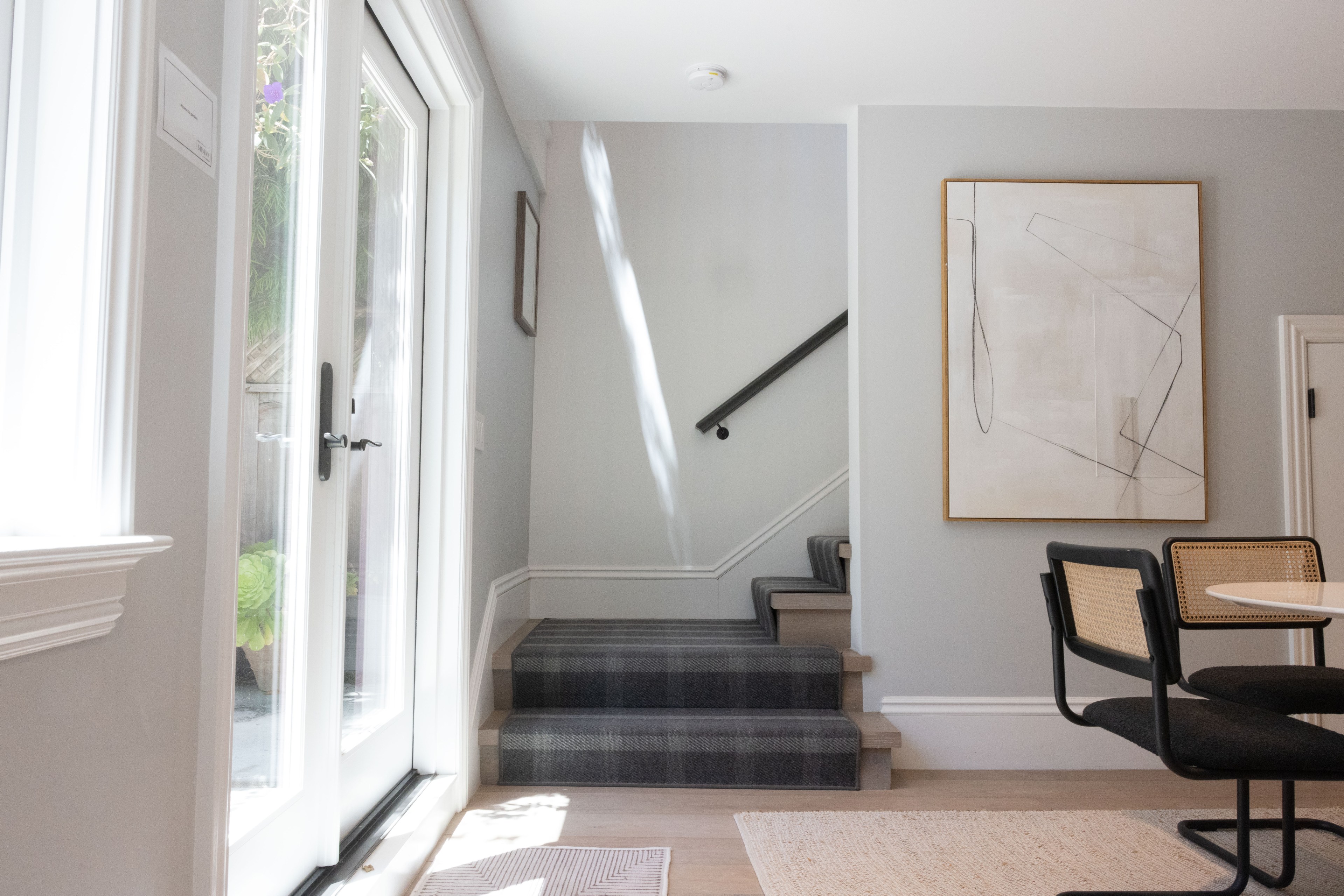 The image shows a well-lit, modern interior with a staircase, a glass door to the outside, abstract wall art, and a dining area with woven back chairs.