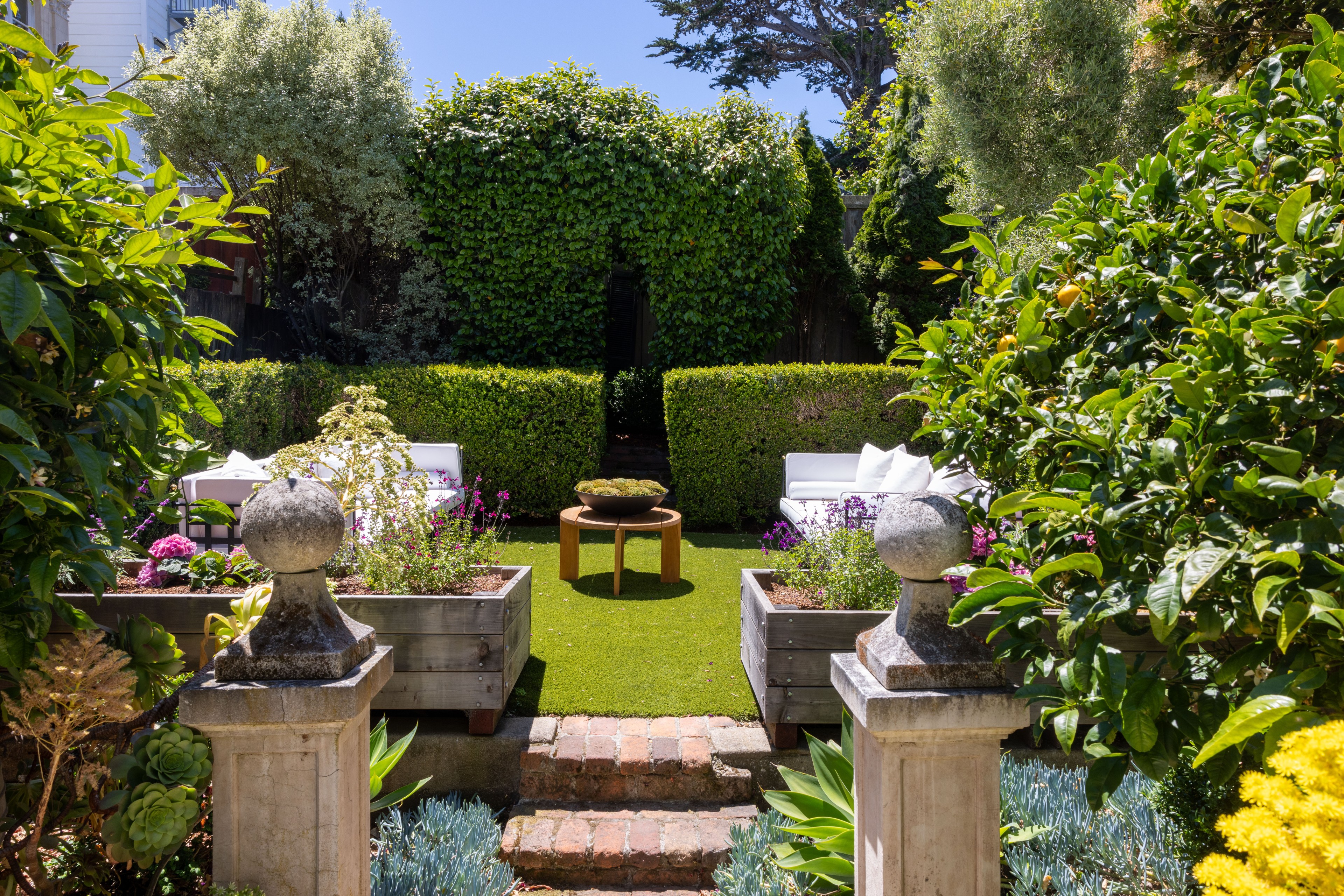 A lush garden features neatly trimmed hedges, colorful flowers, two white cushioned seats, and a central round table. Ornamental plants and stone pillars enhance the scene.