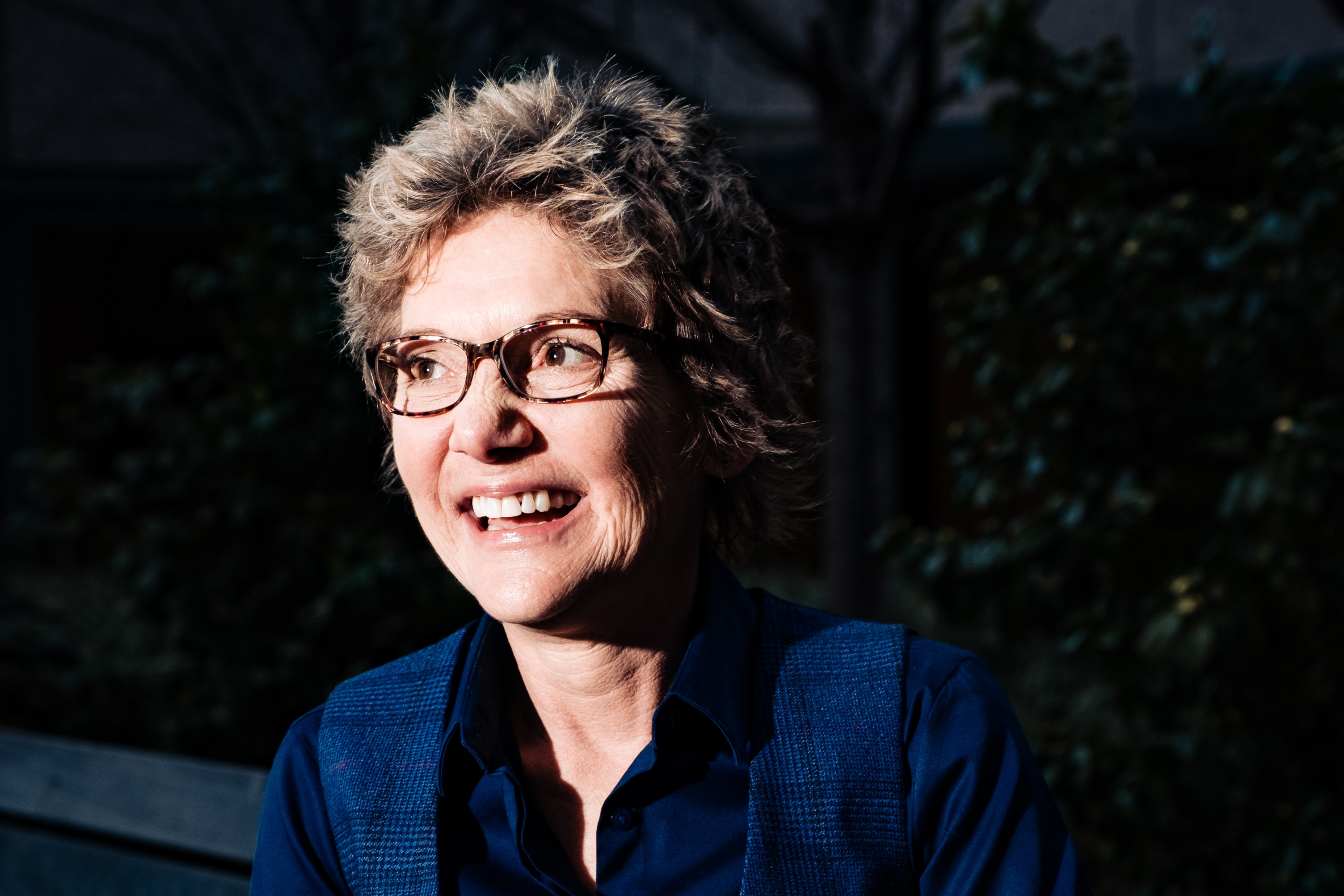 A smiling person with short, wavy hair and glasses is outdoors, wearing a blue shirt and vest, with dark foliage in the background.