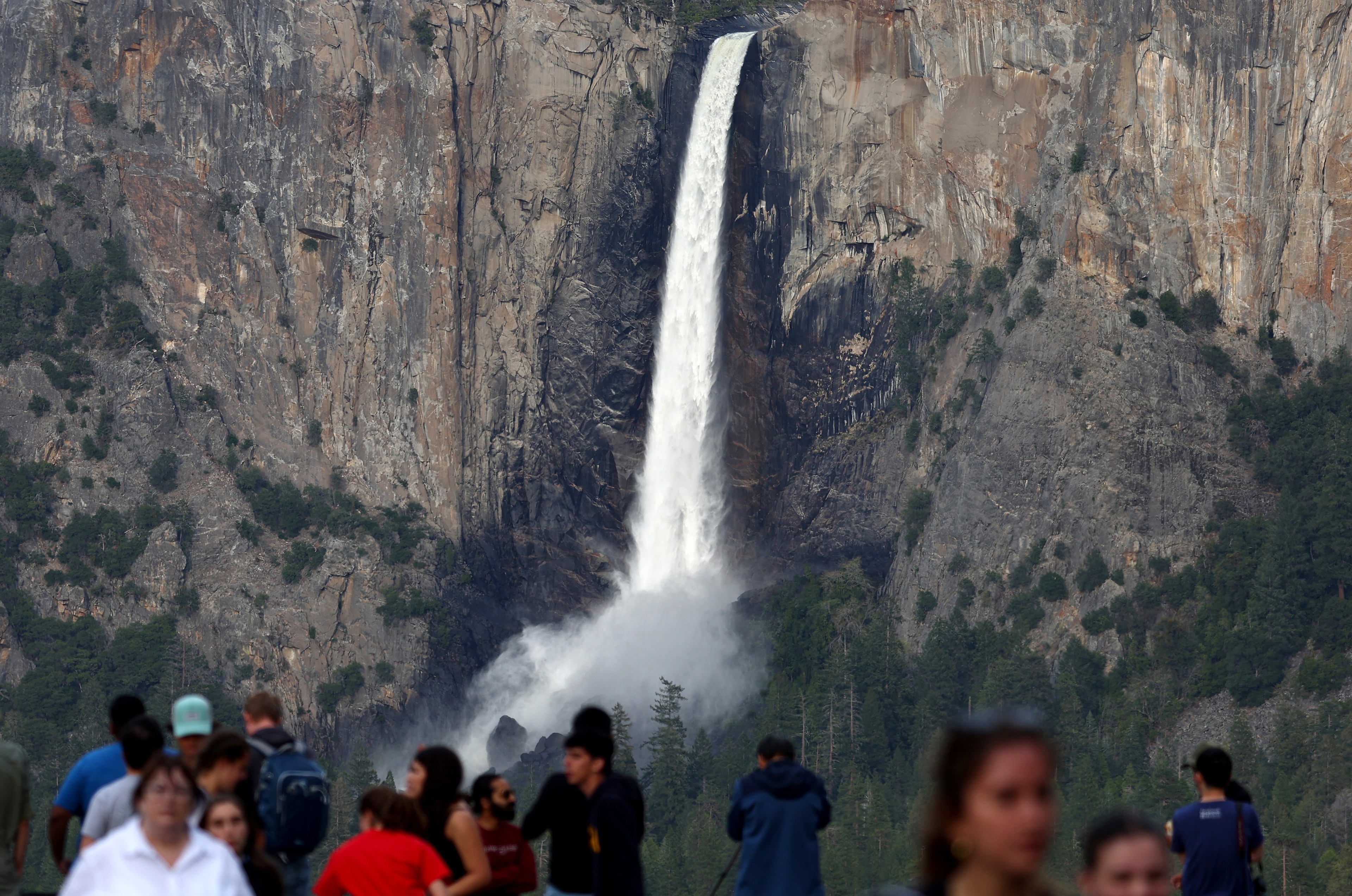 A tall waterfall cascades down a rugged cliffside with mist rising at its base. In the foreground, a crowd of people gathers, partially blurred, observing the scenery.