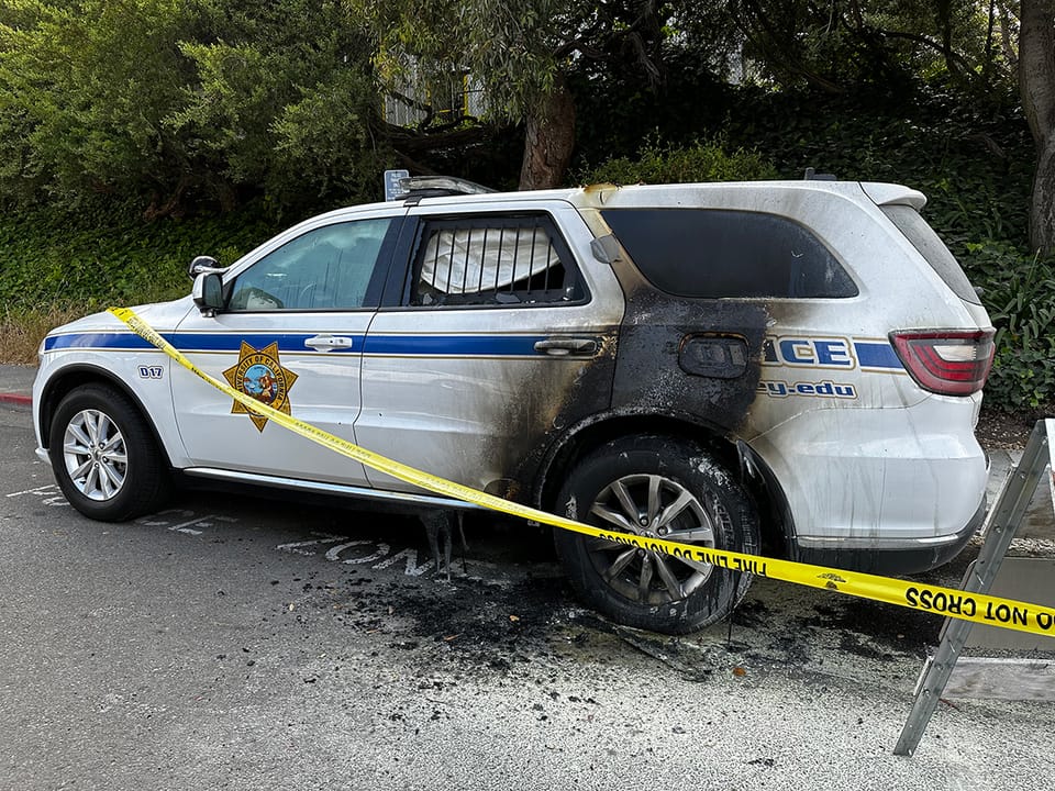 A police SUV with significant fire damage on the rear passenger side is parked beside a curb. Yellow caution tape surrounds the vehicle.