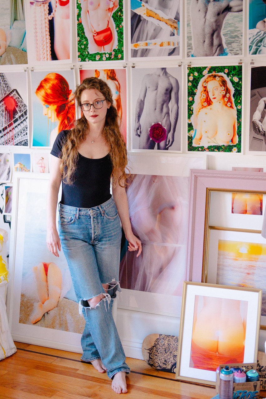 A woman with glasses, wearing a black top and ripped jeans, stands barefoot in front of a wall filled with vibrant photographic prints, mostly of artistic nudes.