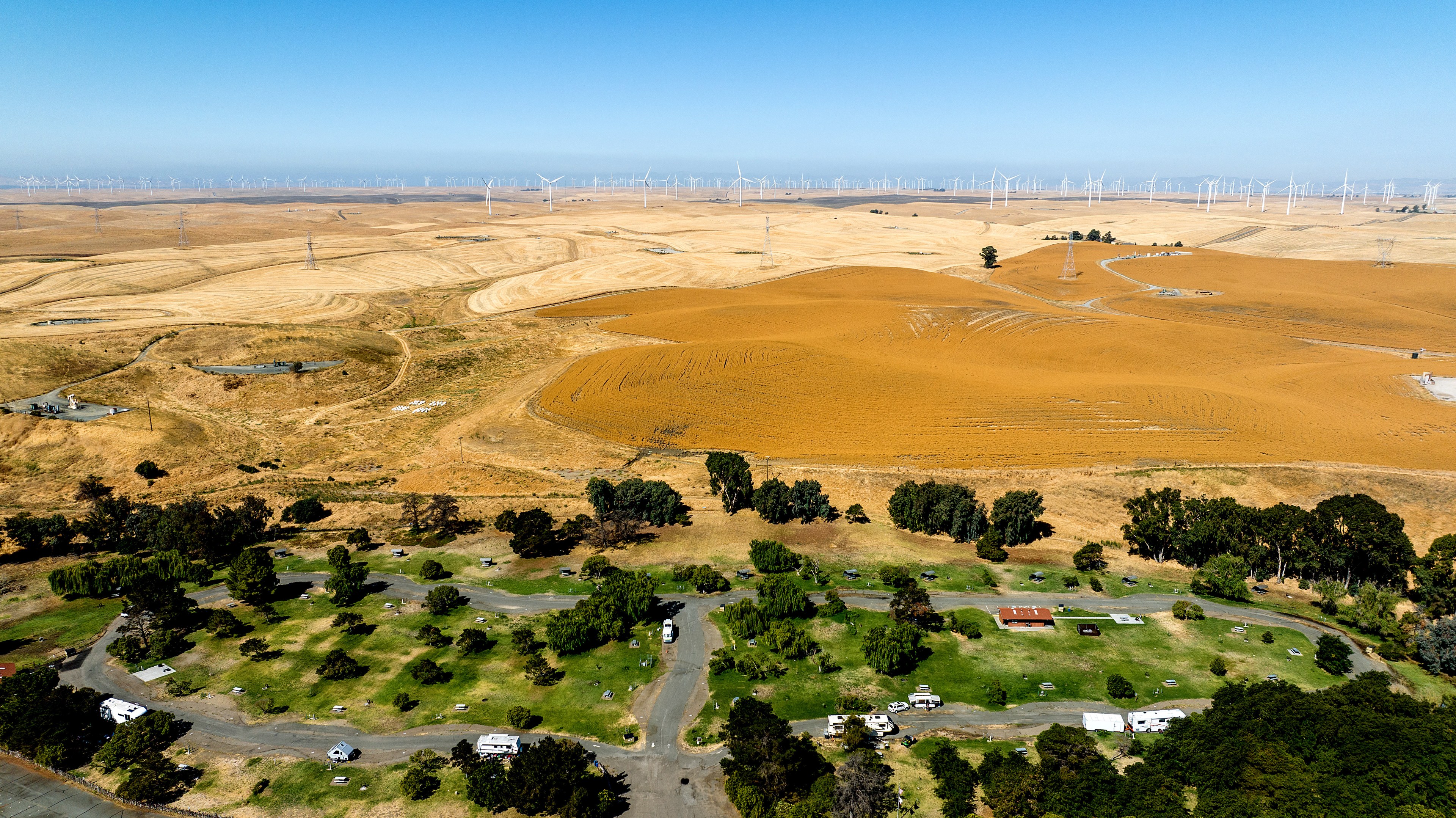 The image features a panoramic view of a hilly landscape with numerous wind turbines in the distance. In the foreground, a RV park is visible with greenery and parked RVs.