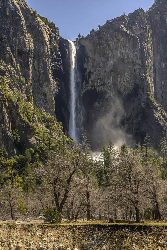 A tall waterfall cascades down a steep rocky cliff surrounded by evergreen trees. Below, leafless trees stand in the foreground under a clear blue sky.