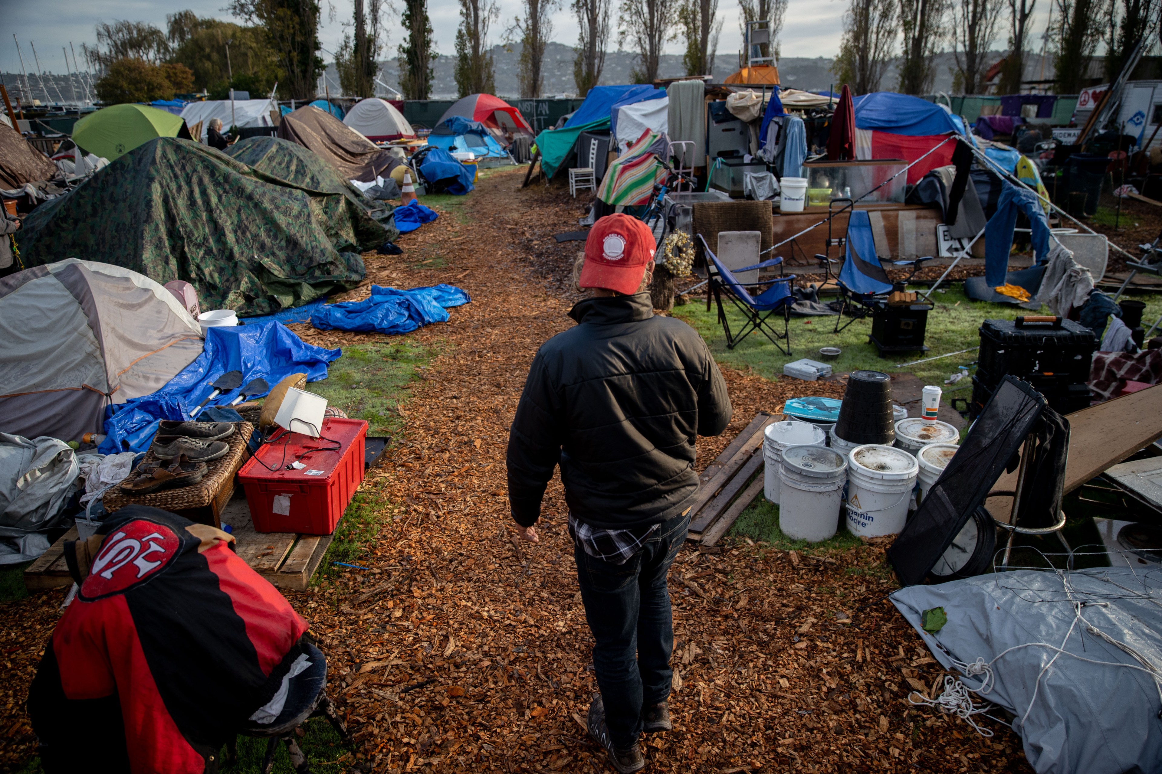 A person wearing a red cap and black jacket walks on a path through a crowded campsite with numerous tents, tarps, and scattered belongings on a grassy field.