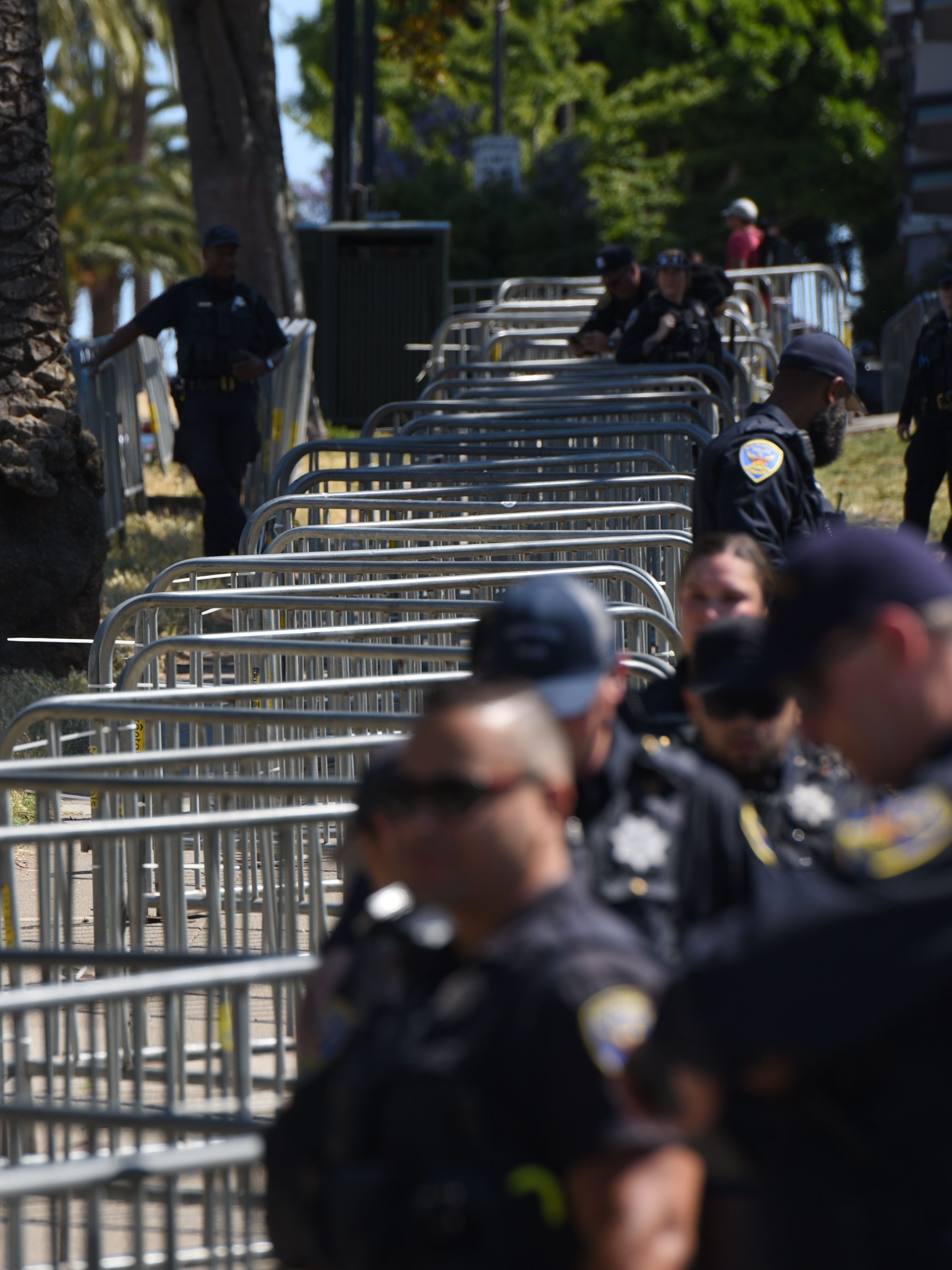 The image shows several police officers standing near metal barricades outdoors, possibly at an event. A parked blue car and trees are visible in the background.