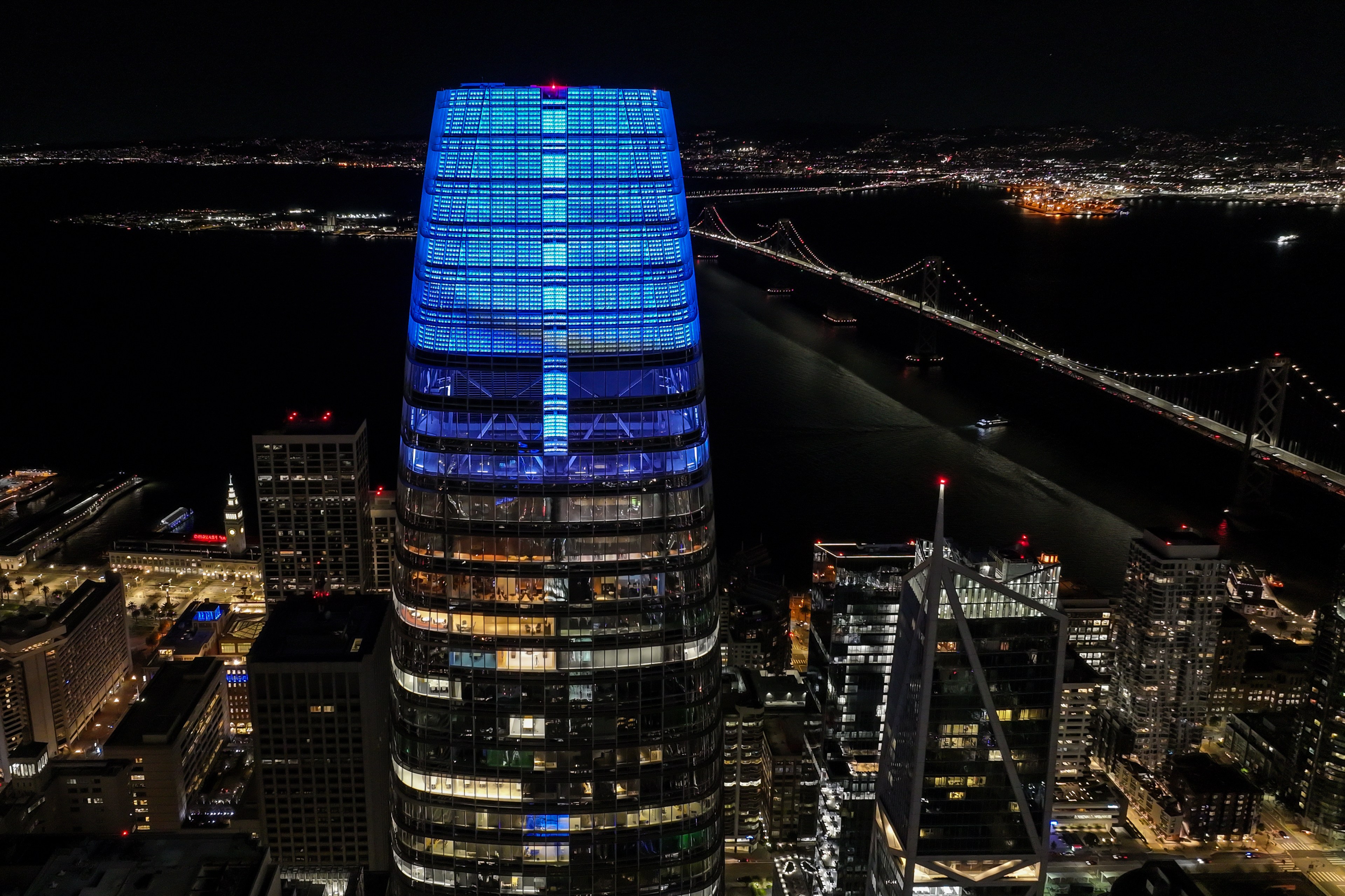 A tall, modern skyscraper topped with blue lights stands prominently in a nighttime cityscape, with a lit suspension bridge stretching across the water in the background.