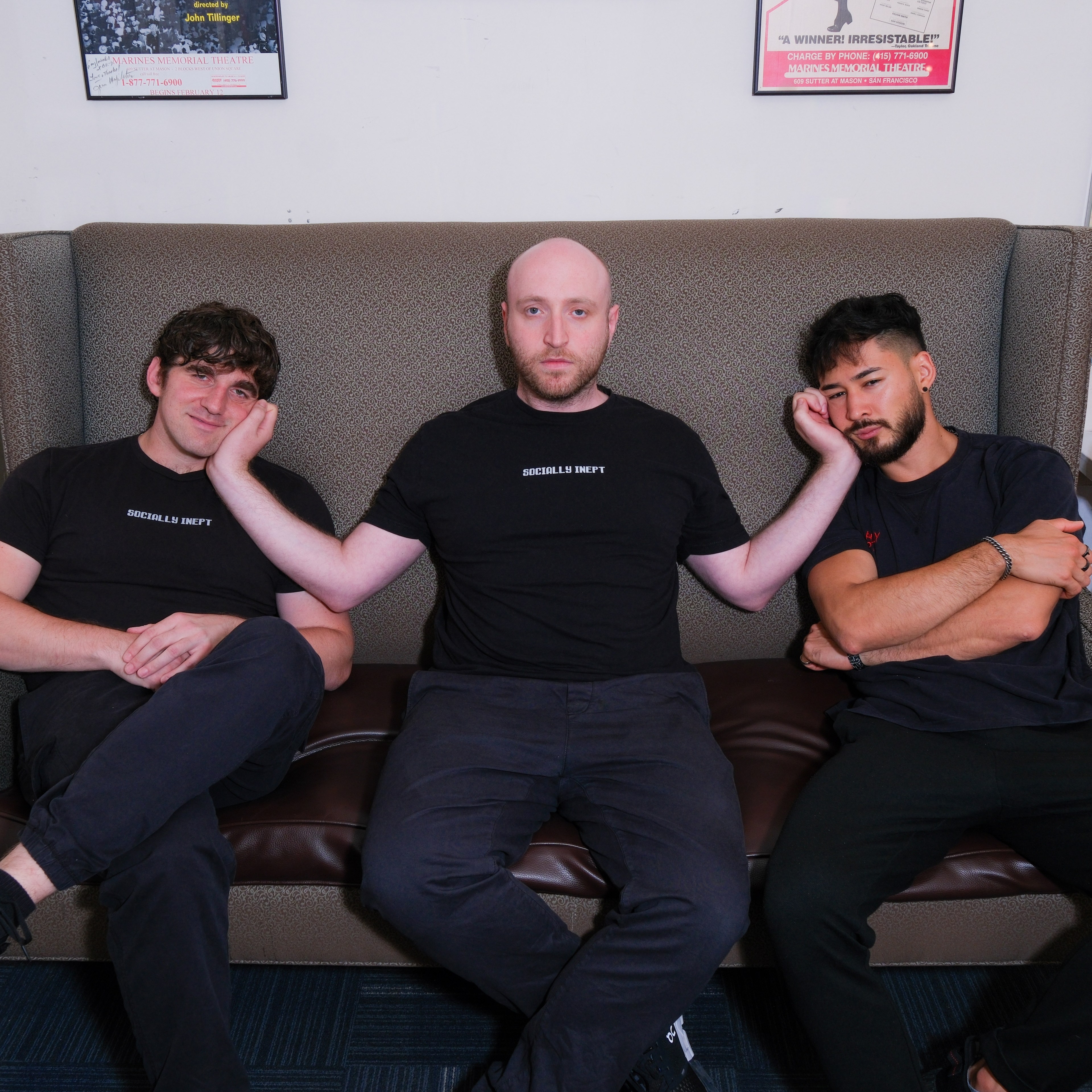 Three men sit on a couch. The man in the middle, wearing a black &quot;SOCIALLY INEPT&quot; shirt, cups the faces of the two men on either side of him, who also wear black.